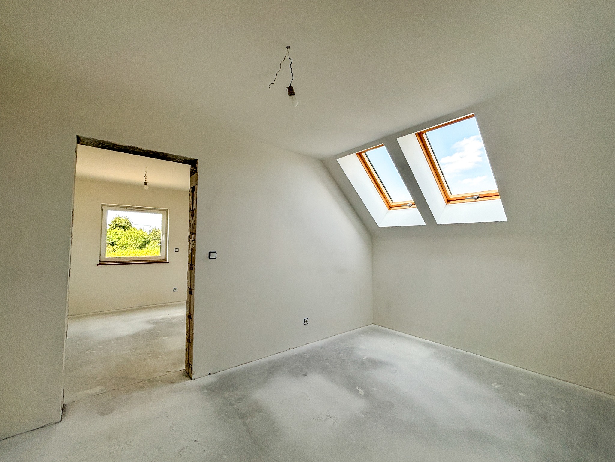 Poddasze w stanie deweloperskim z dwoma oknami dachowymi, świeżo zagruntowane ściany i wylewka podłogowa, widoczne kable od oświetlenia sufitowego i surowe ościeżnice drzwiowe.