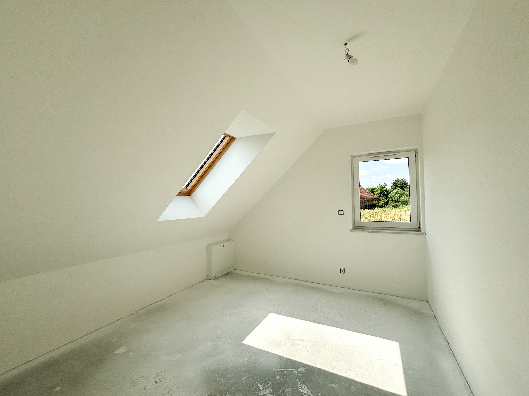 Poddasze w domu jednorodzinnym, po świeżo wykonanych pracach przygotowawczych, z oknem dachowym i widokiem na zewnątrz, surowy beton na podłodze, przygotowany do dalszych prac wykończeniowych.