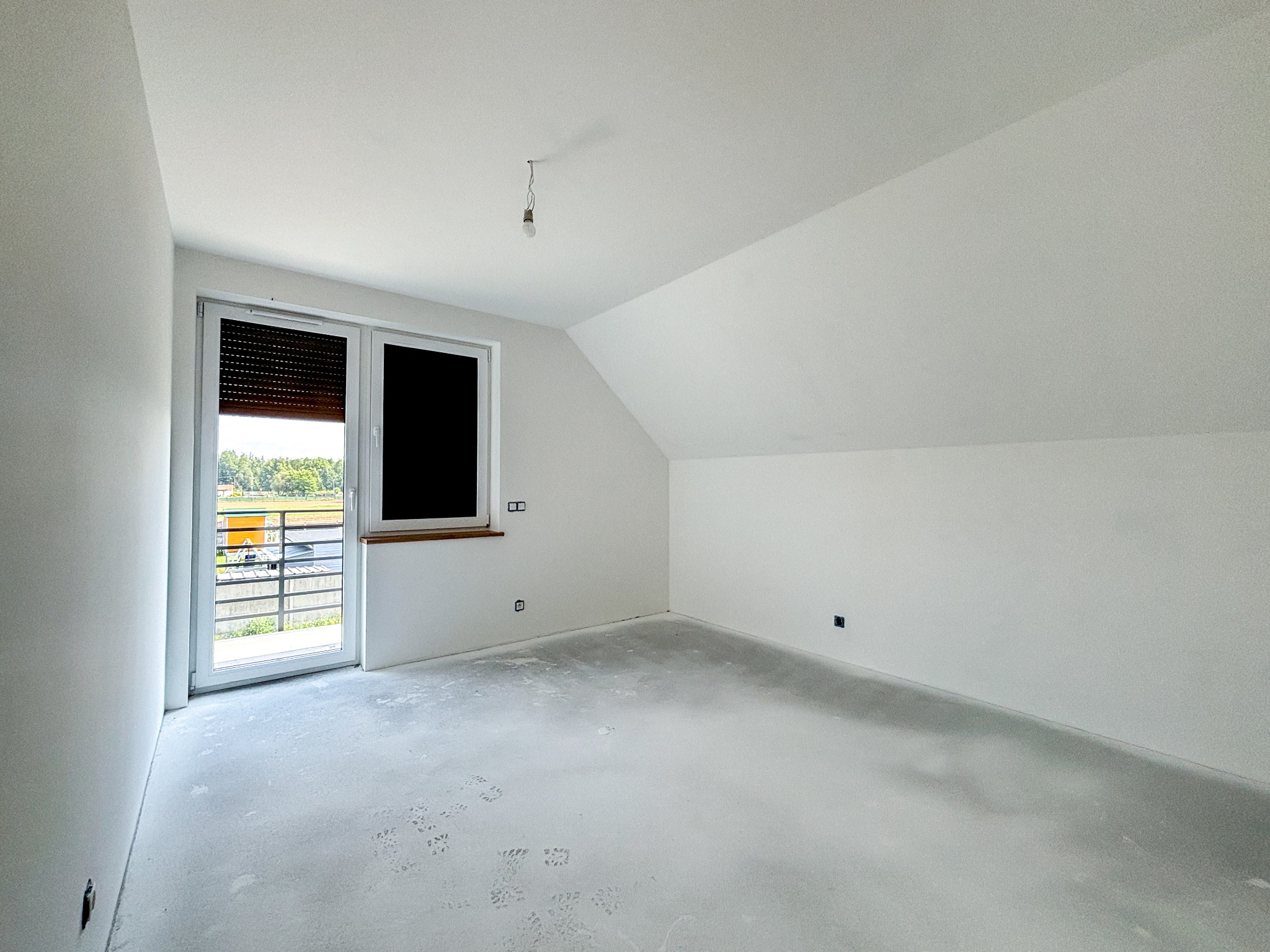 Poddasze w stanie deweloperskim z oknem balkonowym z roletami zewnętrznymi, surowa podłoga, białe ściany i sufit, wisząca żarówka.