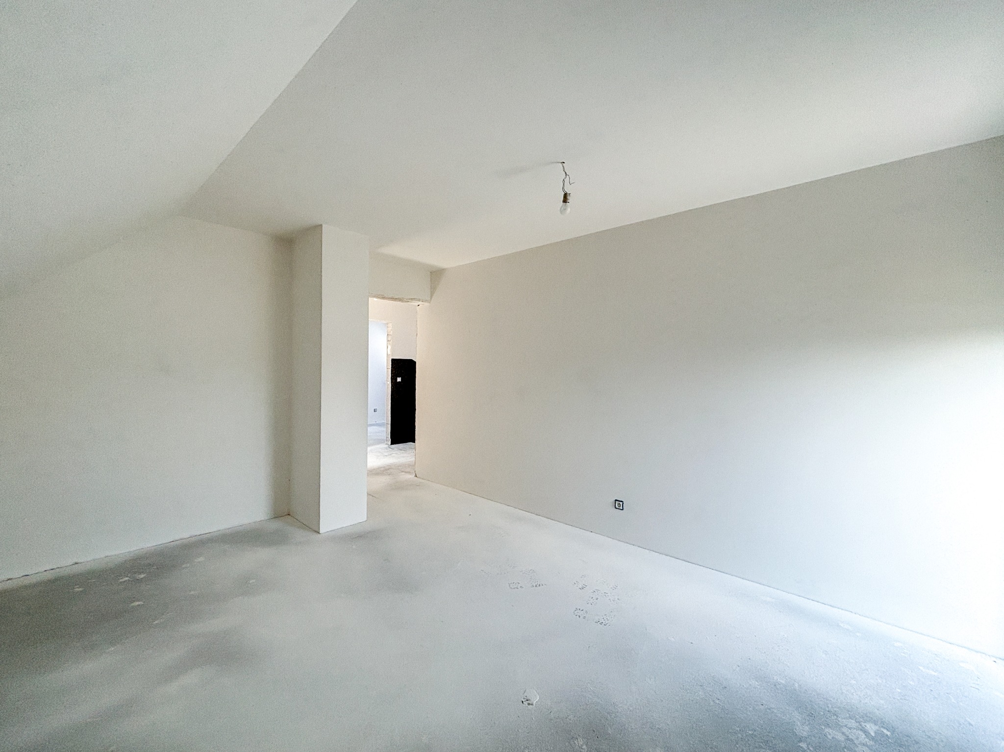 Wnętrze pokoju w trakcie prac wykończeniowych, widoczne świeżo położone gładzie na ścianach i podłodze, surowa żarówka zwisająca z sufitu, widok na sąsiednie pomieszczenie przez otwarte drzwi.