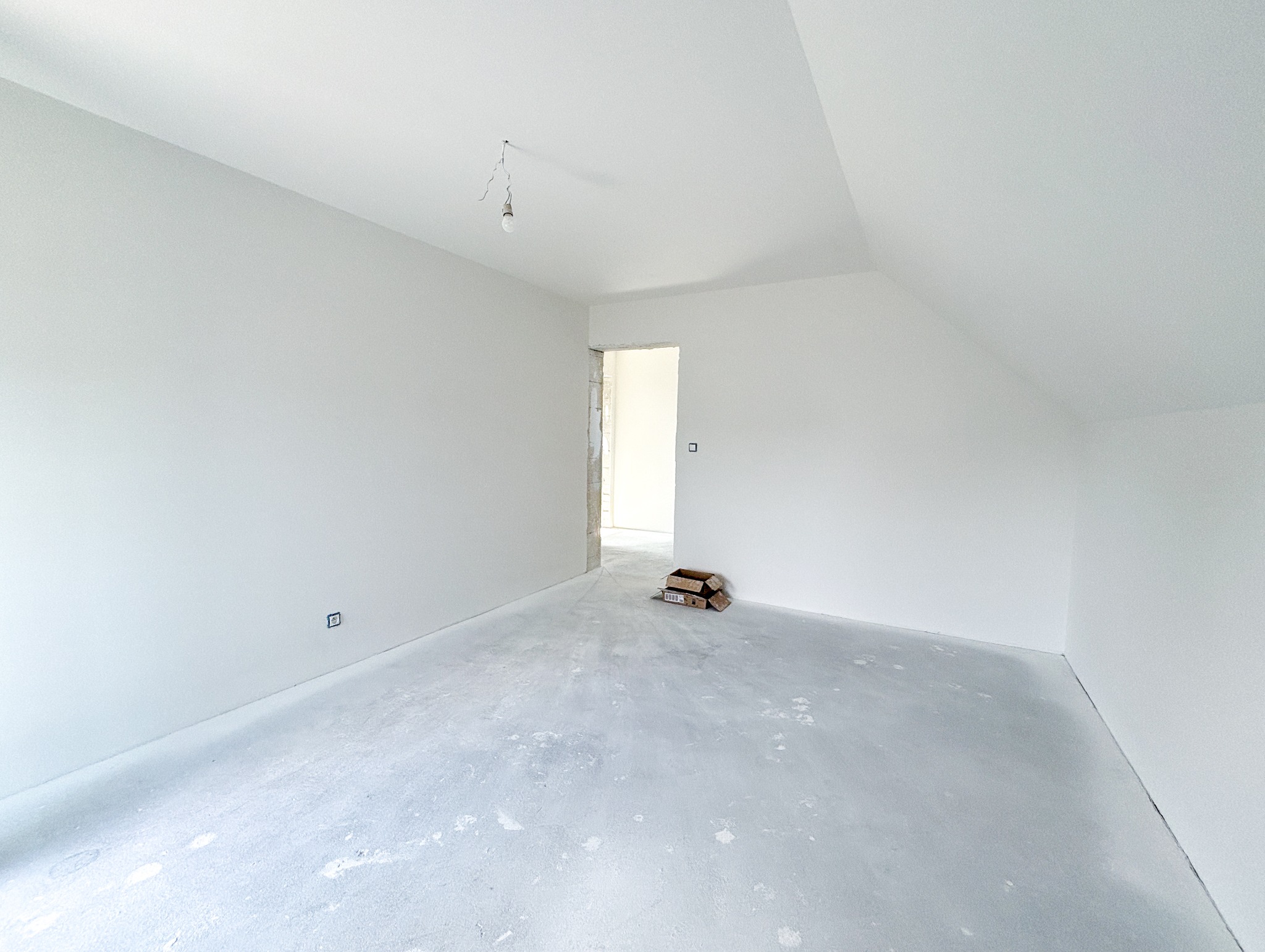 Pusty pokój na poddaszu w trakcie prac wykończeniowych, ściany pomalowane na biało, podłoga z wylewką, widoczne puste kartony i wisząca żarówka.