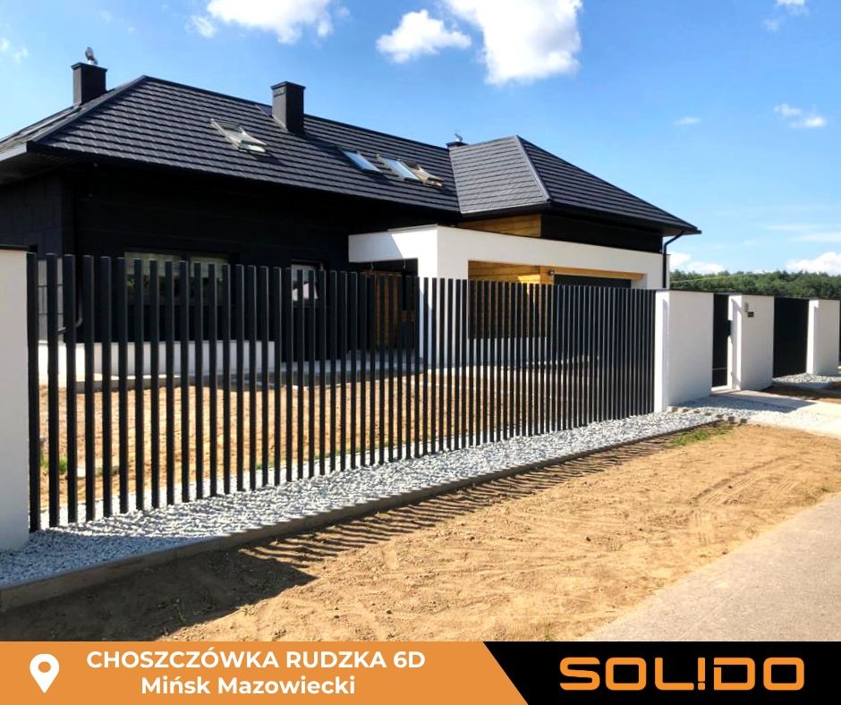 Nowoczesne ogrodzenie panelowe z czarnych pionowych sztachet i białych słupków na tle domu z czarną dachówką i jasną elewacją, widok z zewnątrz na żwirowe podsypanie i piaszczyste podłoże.