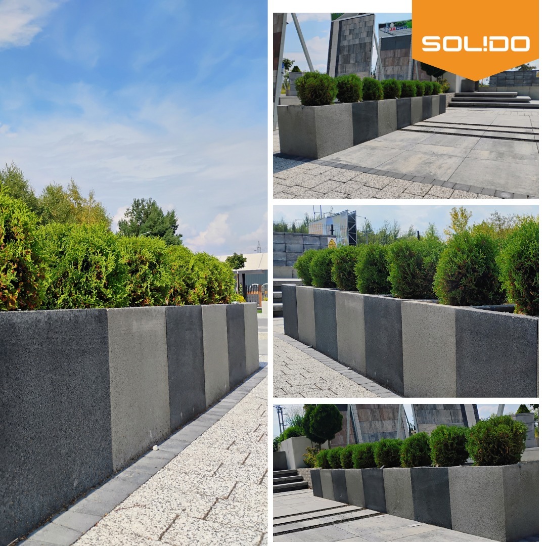 Długi szereg betonowych donic w różnych odcieniach szarości, obsadzonych kulistymi krzewami, tworzących element dekoracyjny w przestrzeni miejskiej. Widoczny chodnik z kostki brukowej i fragment...