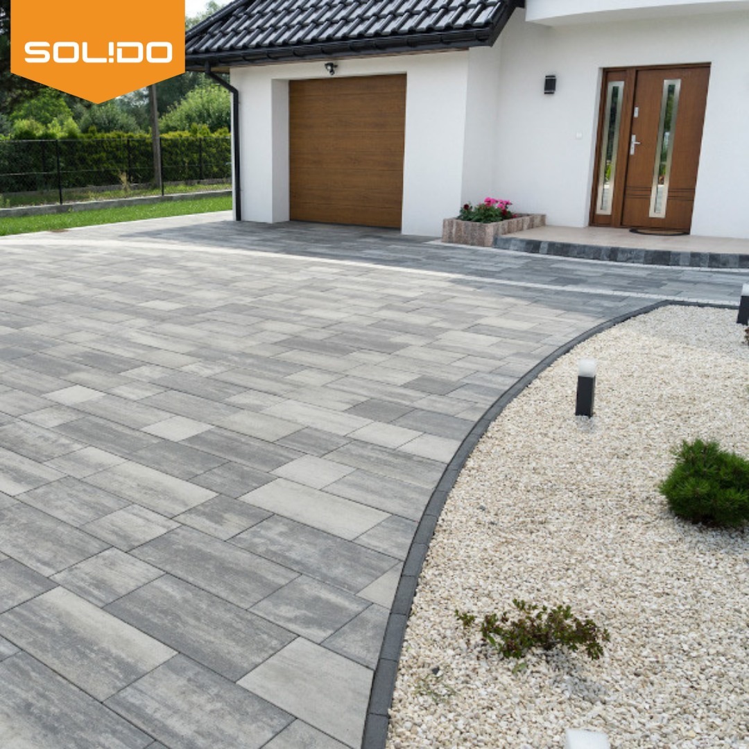 Ułożona szara kostka brukowa przed domem w Choszczówce Rudzkiej, z obrzeżem oddzielającym ją od białego żwiru i niskich krzewów, widoczne drzwi garażowe i wejściowe do domu.