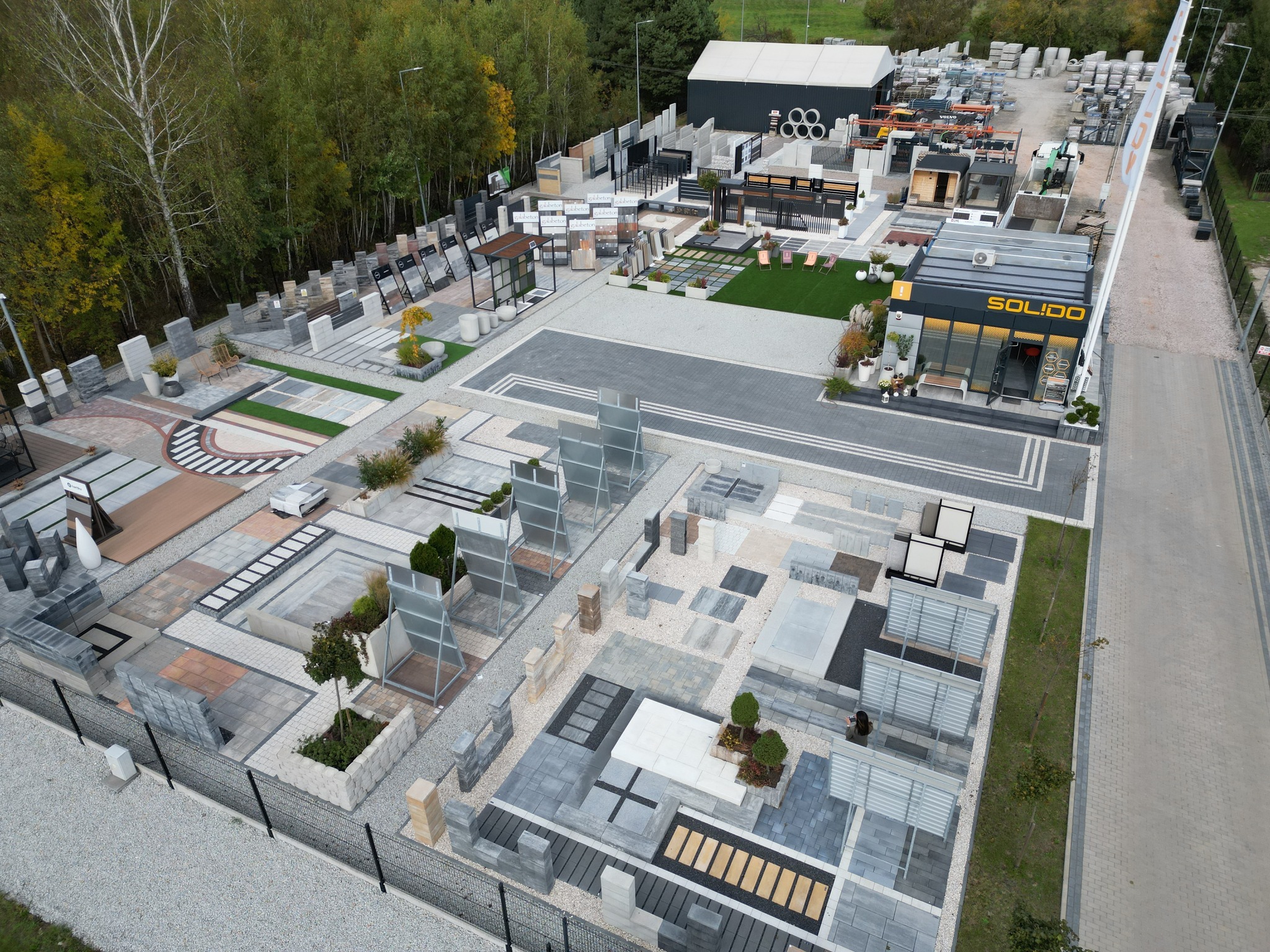 Ekspozycja zewnętrzna kostki brukowej i elementów betonowych, widok z góry, prezentująca różnorodność wzorów i kolorów, otoczona zielenią drzew i krzewów.