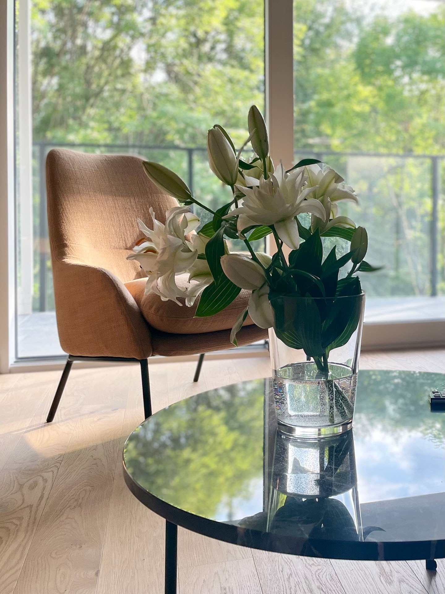 Jasny fotel z beżową tapicerką na tle okna z widokiem na zieleń, stolik kawowy z odbijającym się otoczeniem, w wazonie białe lilie z zielonymi liśćmi.