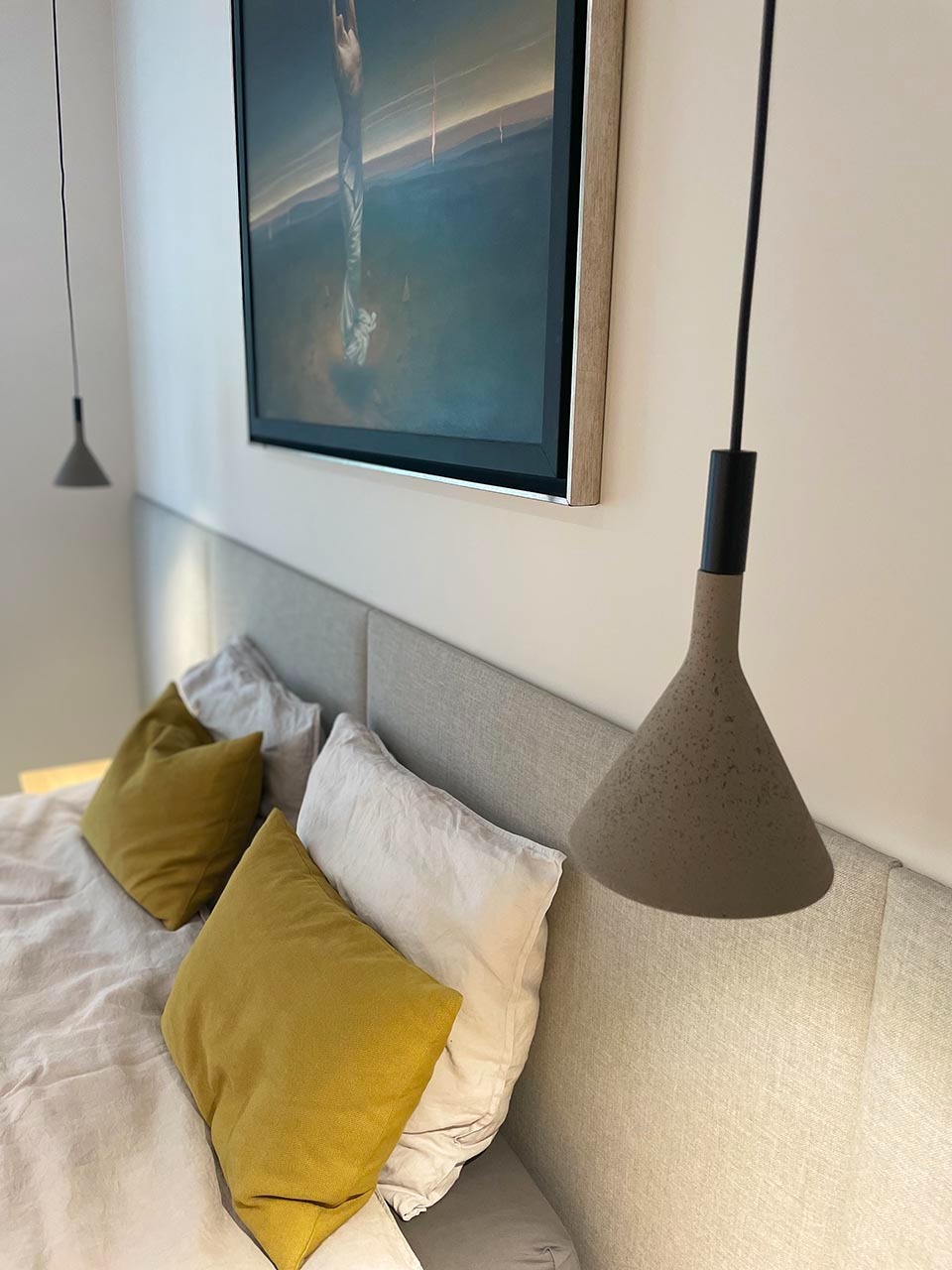 Nowoczesna sypialnia z szarą tapicerowaną ramą łóżka, żółtymi poduszkami i stożkową lampą wiszącą z betonu architektonicznego, nad łóżkiem wisi obraz w drewnianej ramie.