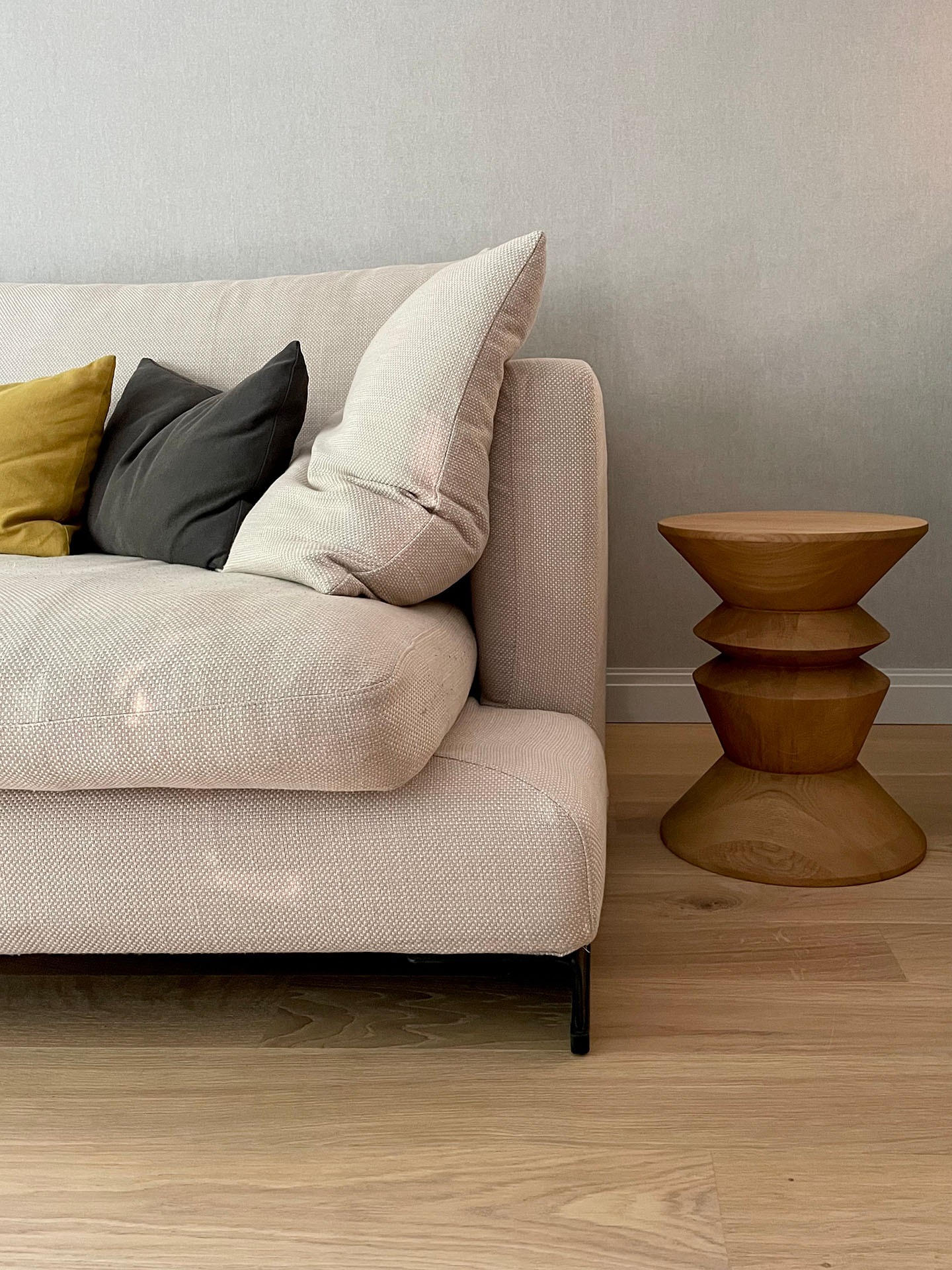 Fragment beżowej sofy z poduszkami w odcieniach żółci i szarości, obok drewniany stolik o kształcie klepsydry na jasnej drewnianej podłodze.