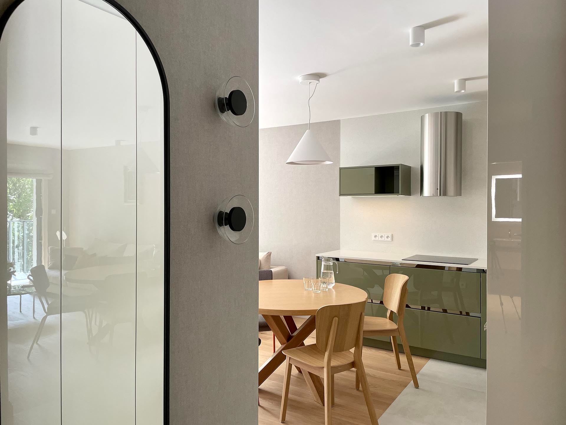 Nowoczesna kuchnia z okrągłym drewnianym stołem i krzesłami, widziana przez przeszklone drzwi z czarną ramą, z widocznymi uchwytami na ścianie i minimalistycznym oświetleniem.