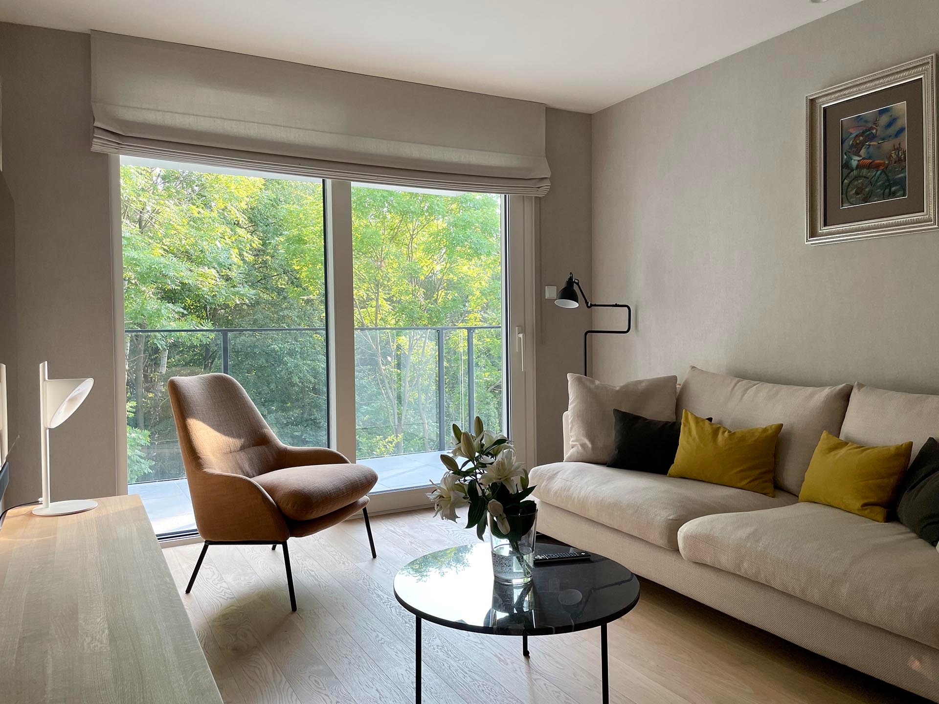 Jasny salon z beżową sofą, fotel w odcieniach brązu, okrągły stolik kawowy z czarnym blatem i widokiem na zieleń za oknem balkonowym.