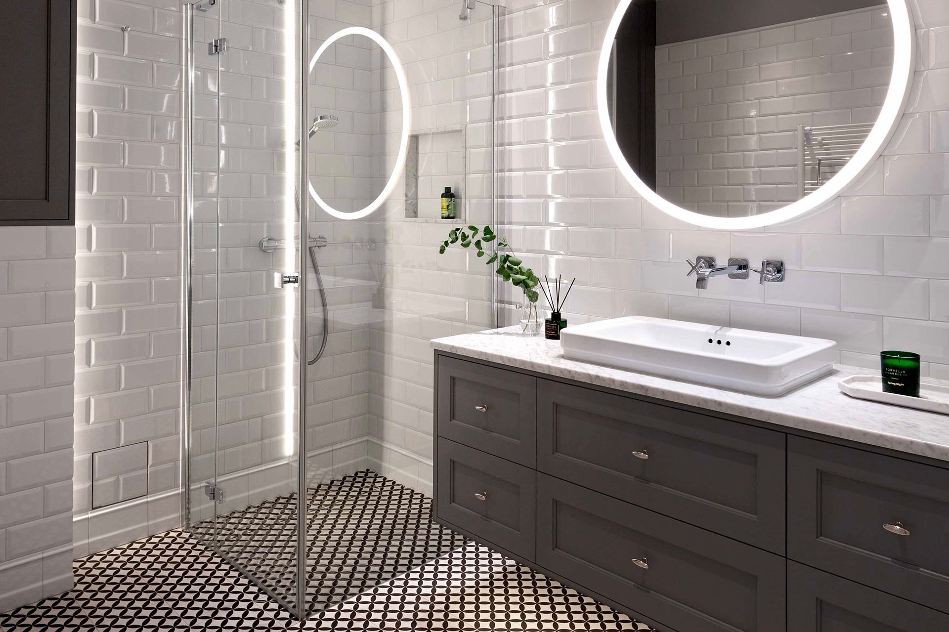 Nowoczesna łazienka z białą cegłą, okrągłymi lustrami z podświetleniem LED, kabiną prysznicową ze szklaną ścianką i wzorzystą czarno-białą podłogą.