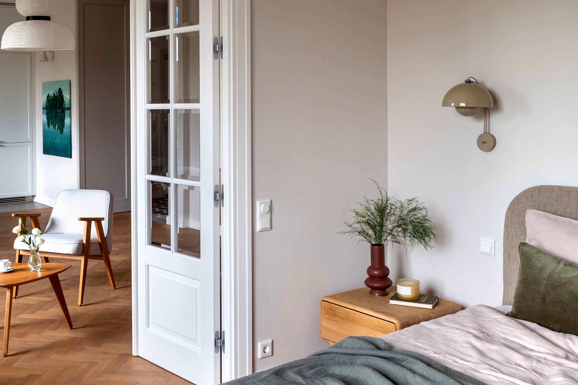 Jasna sypialnia z drewnianą podłogą w jodełkę, łóżkiem z beżowym zagłówkiem, zieloną pościelą i stolikiem nocnym z wazonem i świecą, widok na salon przez białe, przeszklone drzwi.