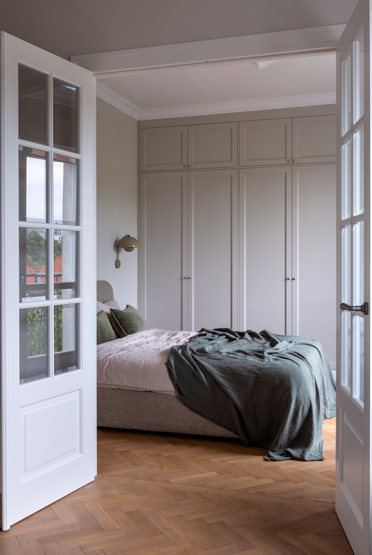 Sypialnia z parkietem w jodełkę, szarą zabudową szaf, łóżkiem z zielonym pledem i otwartymi białymi drzwiami.