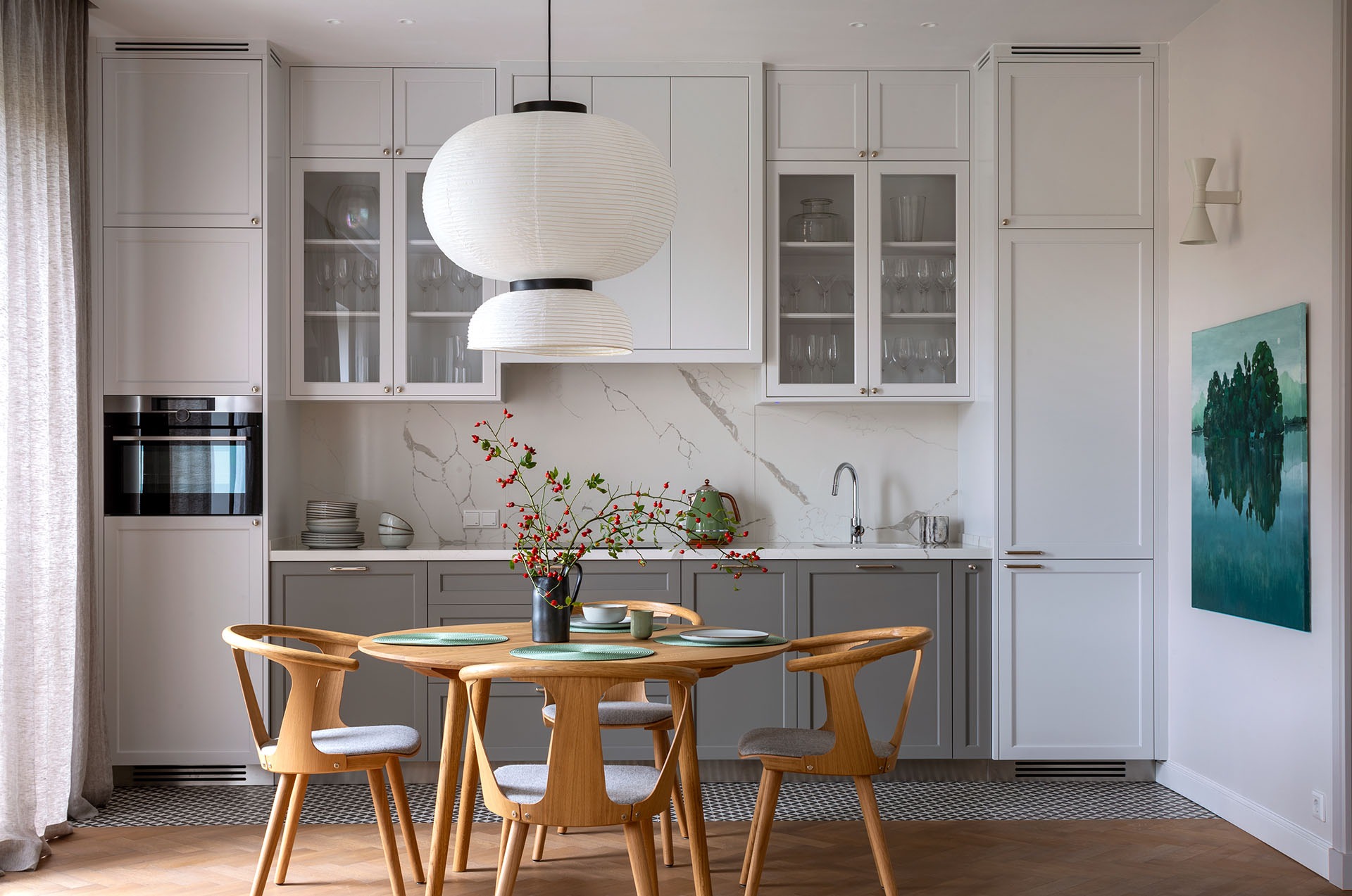 Jasna kuchnia z drewnianym stołem i krzesłami, nad stołem wisi duża papierowa lampa, na ścianie obraz z motywem leśnym, widoczne szklane szafki i sprzęt AGD.
