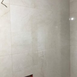 MAL-BUD - Ściana wyłożona jasnymi, marmurkowymi płytkami. Widoczna puszka elektryczna z przewodami oraz haczyk na ścianie.