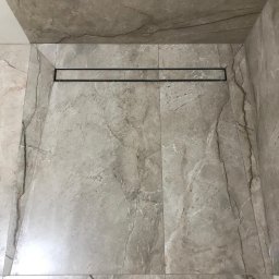 MAL-BUD - Ukończona kabina prysznicowa walk-in z odpływem liniowym, wyłożona szarymi płytkami imitującymi marmur, widoczne fragmenty butów pracownika w dolnej części kadru.