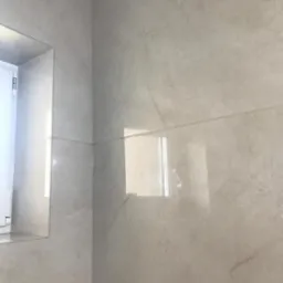 Ściana i narożnik łazienki wyłożone jasnoszarymi płytkami imitującymi kamień, z odbiciem okna w jednej z płytek.