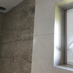 MAL-BUD - Wykończona ściana łazienki z kombinacją płytek imitujących marmur w odcieniach szarości i beżu, obok okna z widocznym fragmentem grzejnika.