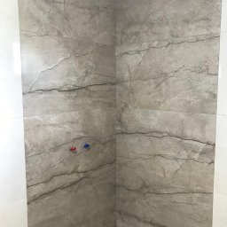 MAL-BUD - Ściany kabiny prysznicowej wyłożone płytkami imitującymi marmur, widoczne przyłącza wody z zaślepkami w kolorze czerwonym i niebieskim.