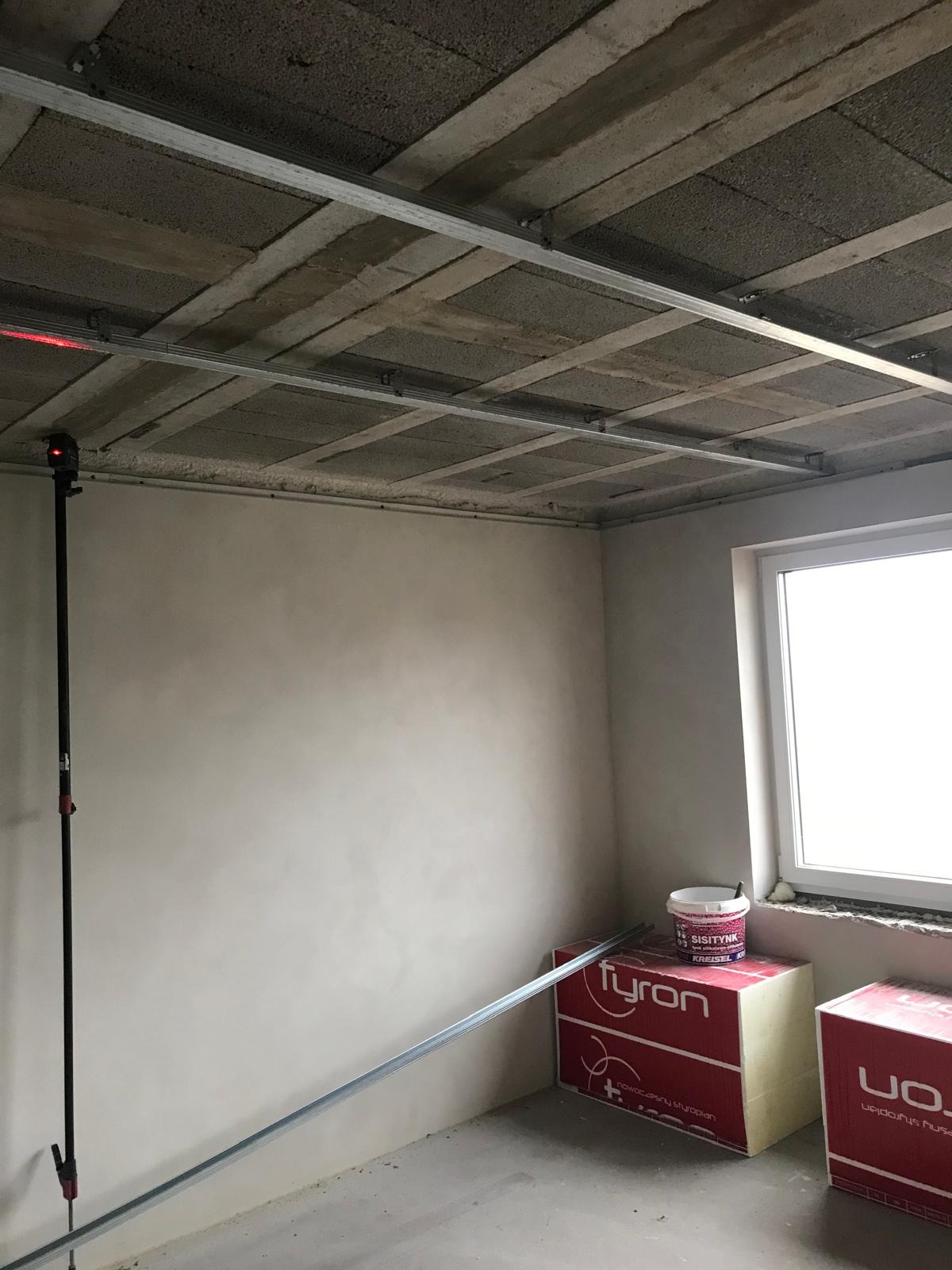 Surowy stan pokoju z zamontowanym stelażem sufitu podwieszanego, widoczny laser krzyżowy na statywie, ściany przygotowane do malowania, materiały budowlane przy oknie.