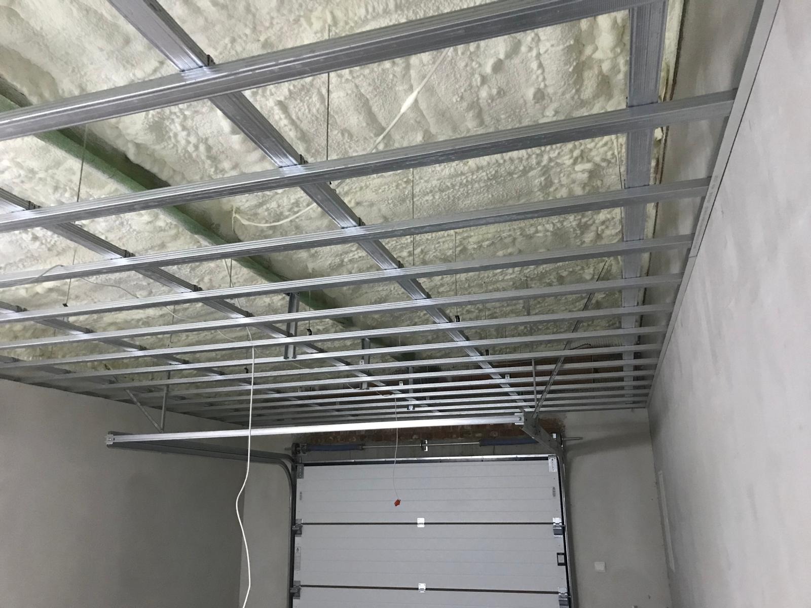 Sufit w trakcie ocieplania pianką poliuretanową, widoczna metalowa konstrukcja podwieszana, brama garażowa w tle.