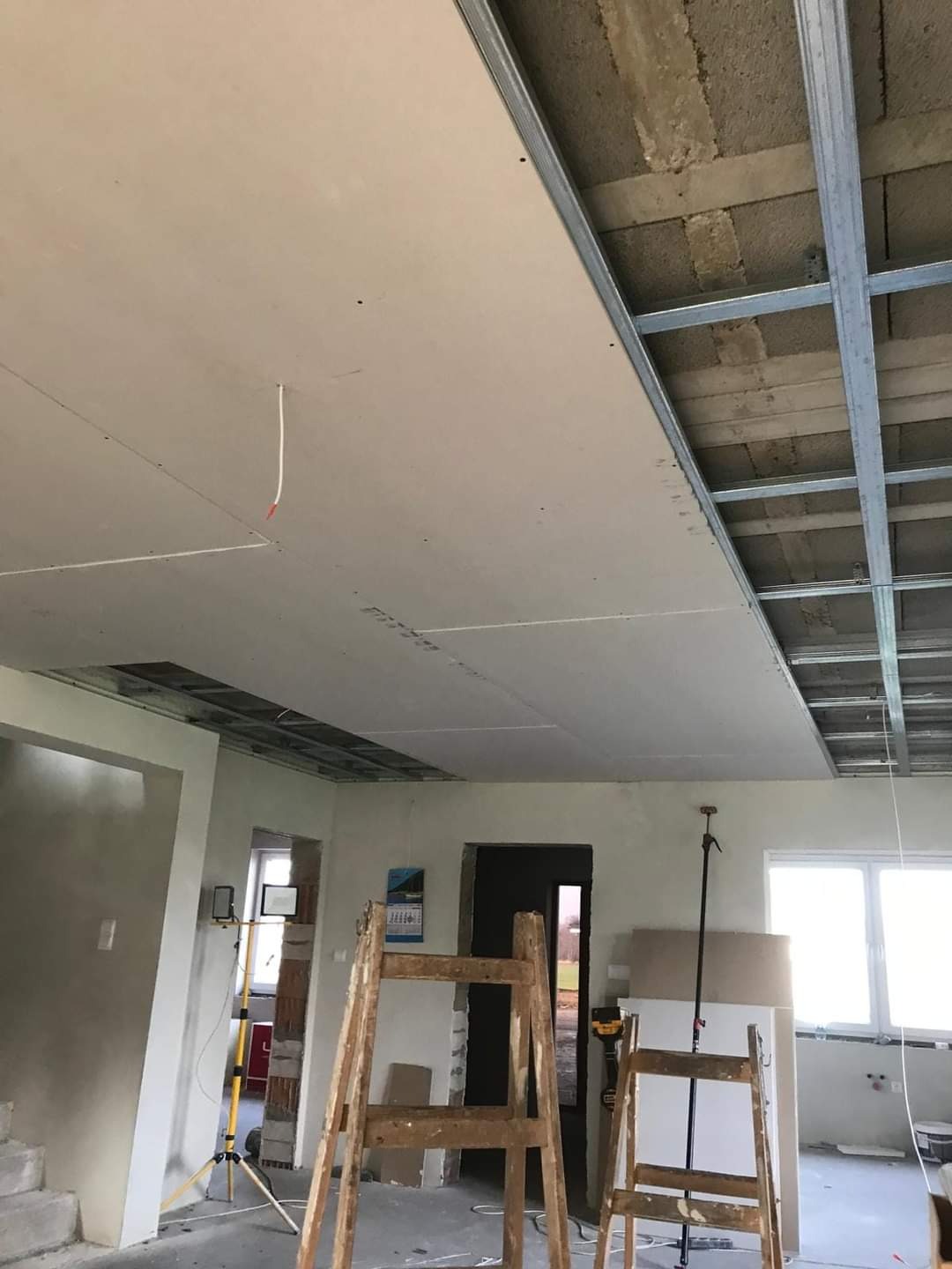 Montaż sufitu podwieszanego z płyt gipsowo-kartonowych na metalowej konstrukcji, widoczne drabiny i narzędzia w surowym wnętrzu domu w trakcie prac wykończeniowych.