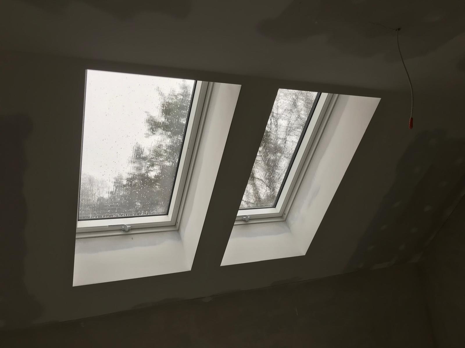 Dwa okna dachowe zamontowane w surowym, szpachlowanym suficie na poddaszu. Przez szyby widać zewnątrz, gdzie pada deszcz na drzewa.