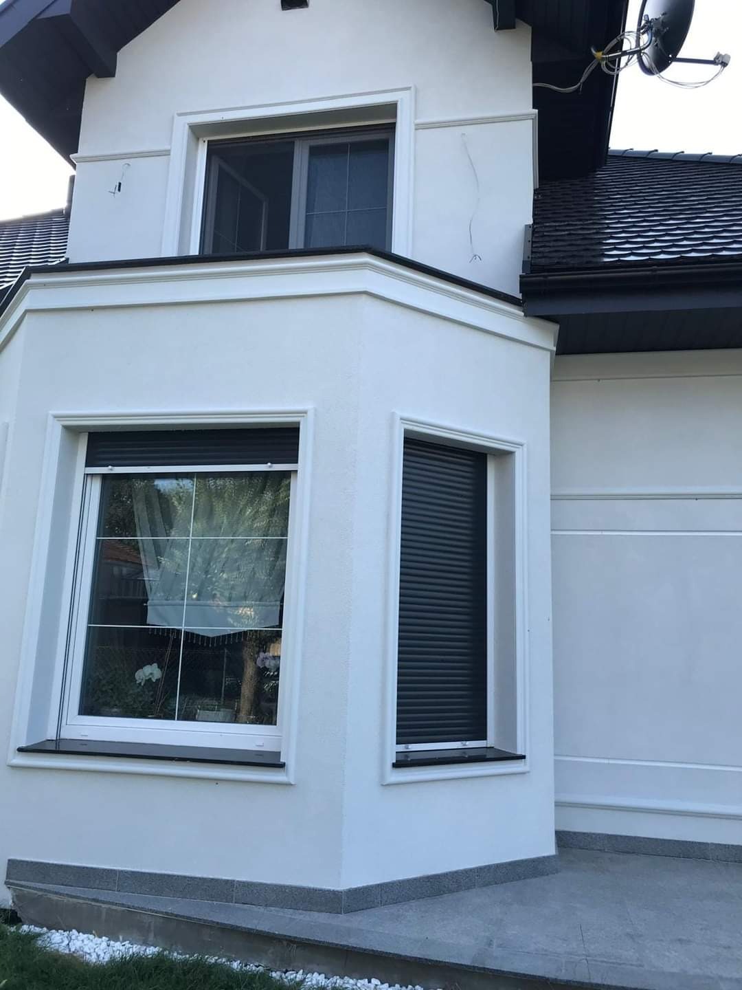 Elewacja domu z białym tynkiem, widoczne okna z roletami zewnętrznymi, jedno okno z firanką, czarny dach z anteną satelitarną.