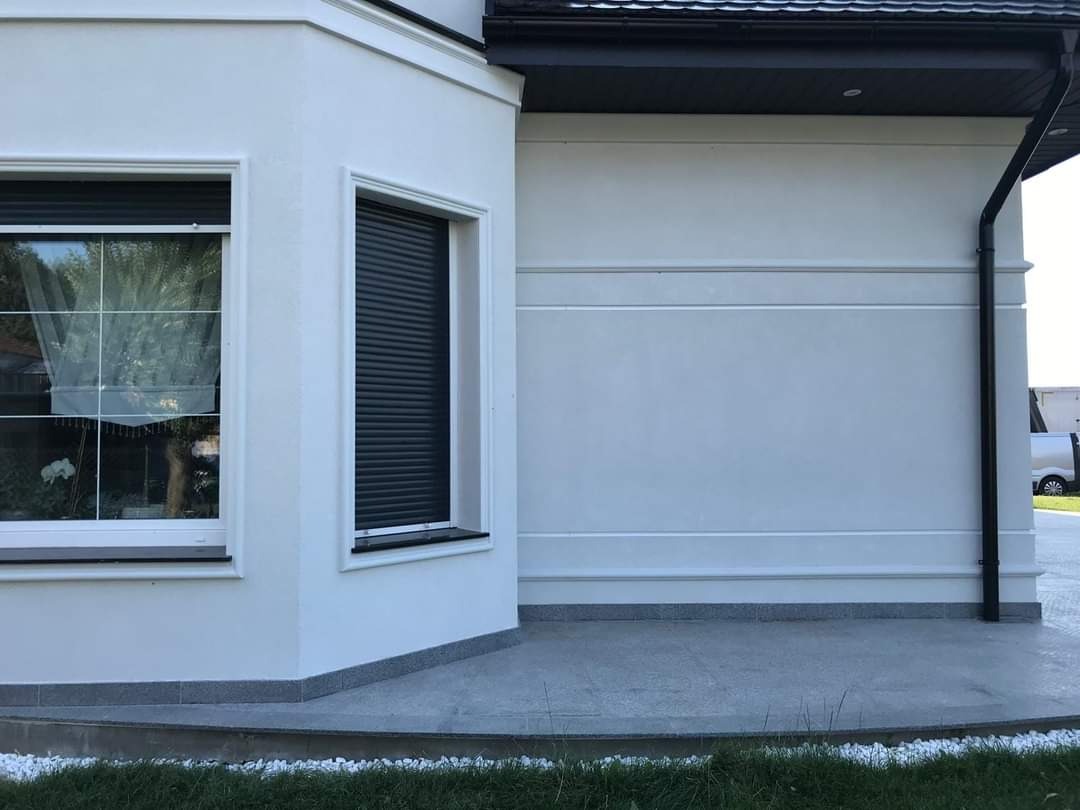 Elewacja budynku z oknami, roletami zewnętrznymi i elementami dekoracyjnymi, widoczny fragment dachu i rynny, trawnik w tle.