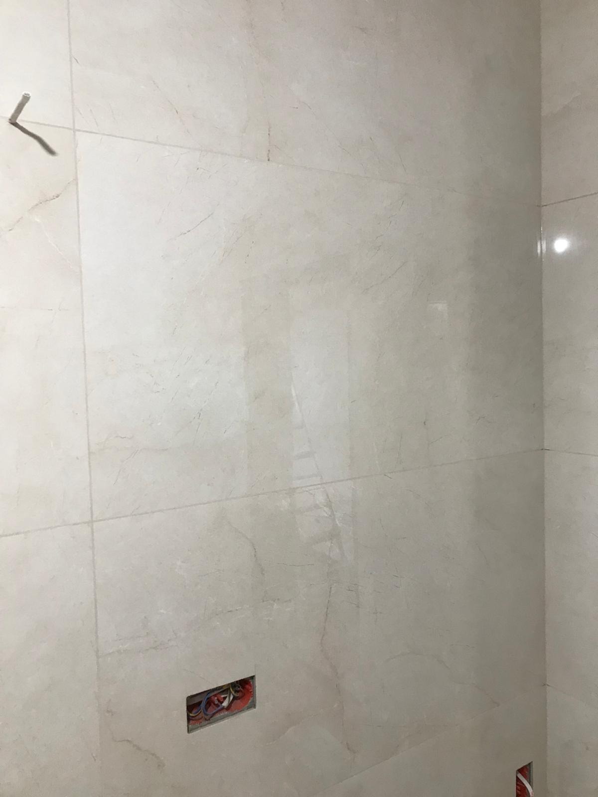 Ściana wyłożona jasnymi, marmurkowymi płytkami. Widoczna puszka elektryczna z przewodami oraz haczyk na ścianie.