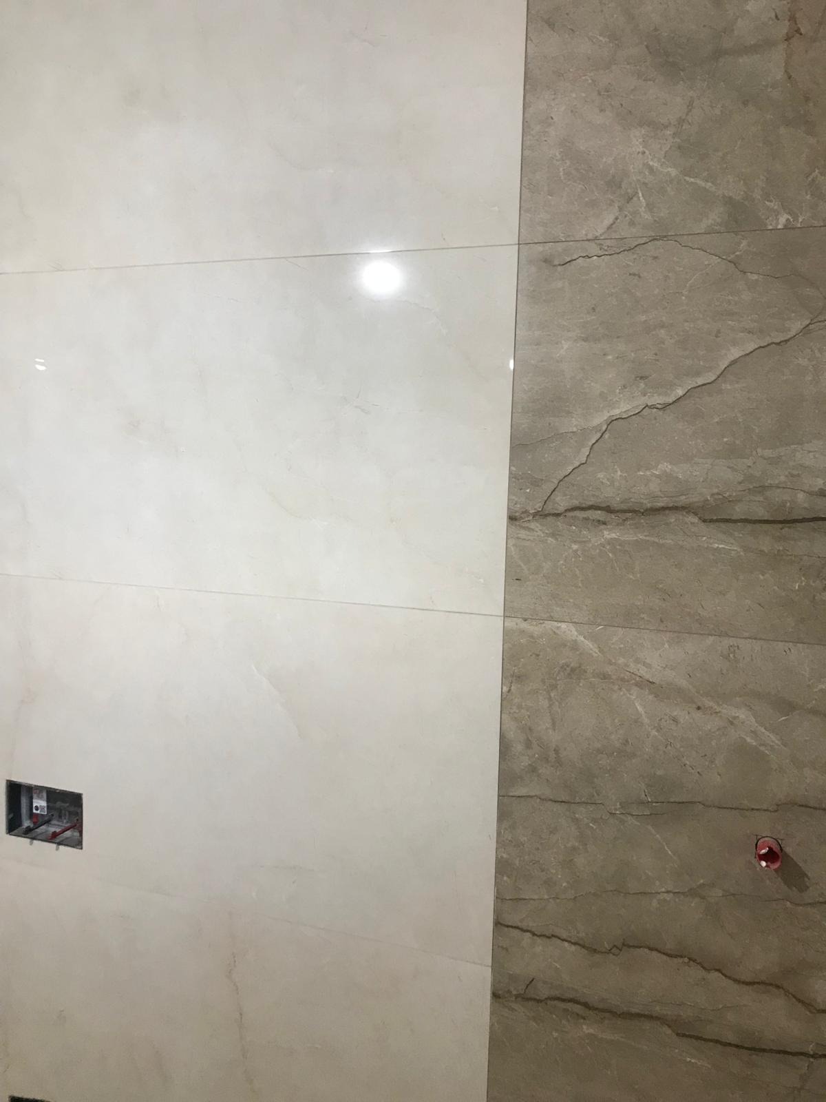 Ściana wyłożona dwoma rodzajami płytek imitujących marmur: jasnokremowe i szare z wyraźnym użyleniem. Widoczna puszka elektryczna bez osprzętu i fragment instalacji hydraulicznej.