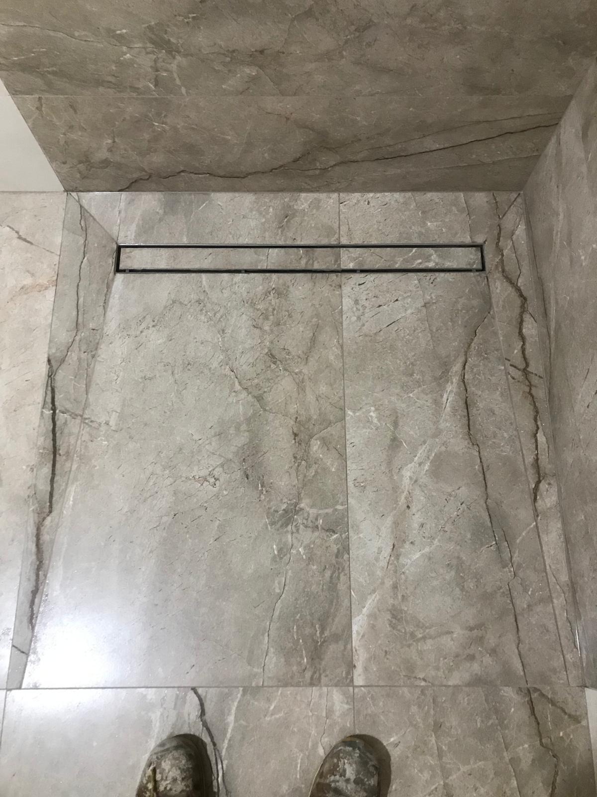 Ukończona kabina prysznicowa walk-in z odpływem liniowym, wyłożona szarymi płytkami imitującymi marmur, widoczne fragmenty butów pracownika w dolnej części kadru.