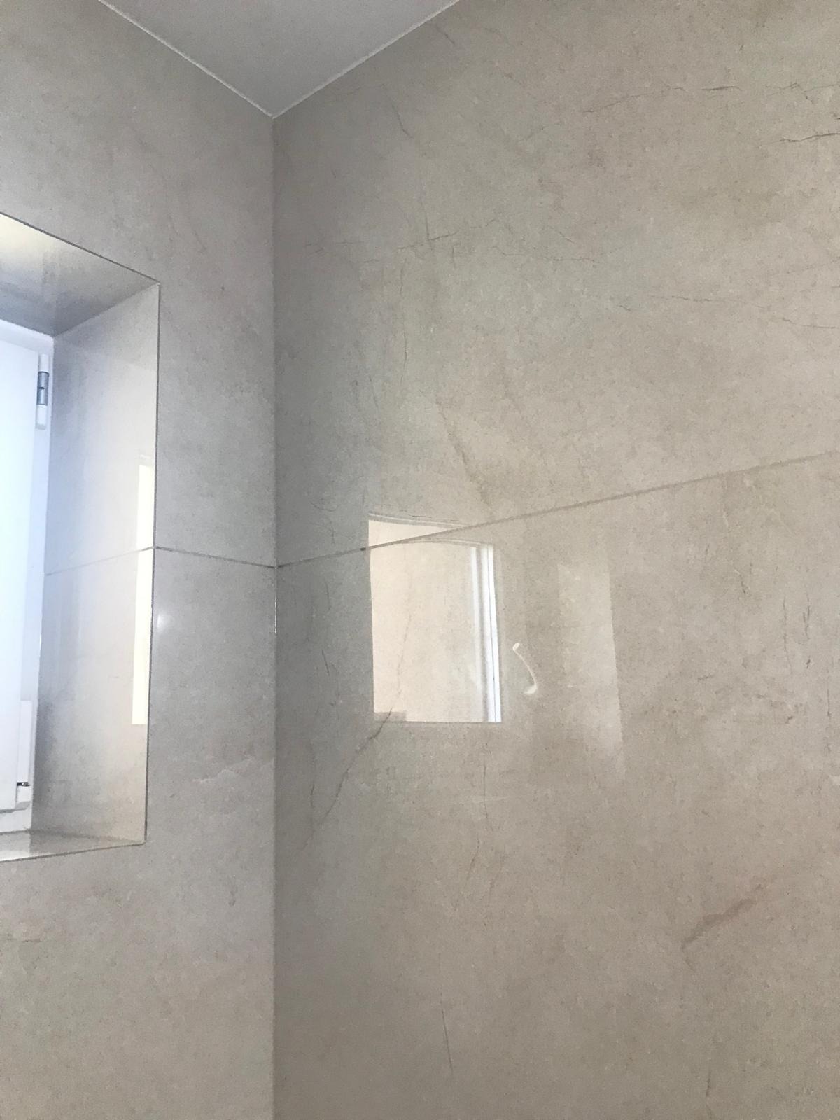 Ściana i narożnik łazienki wyłożone jasnoszarymi płytkami imitującymi kamień, z odbiciem okna w jednej z płytek.