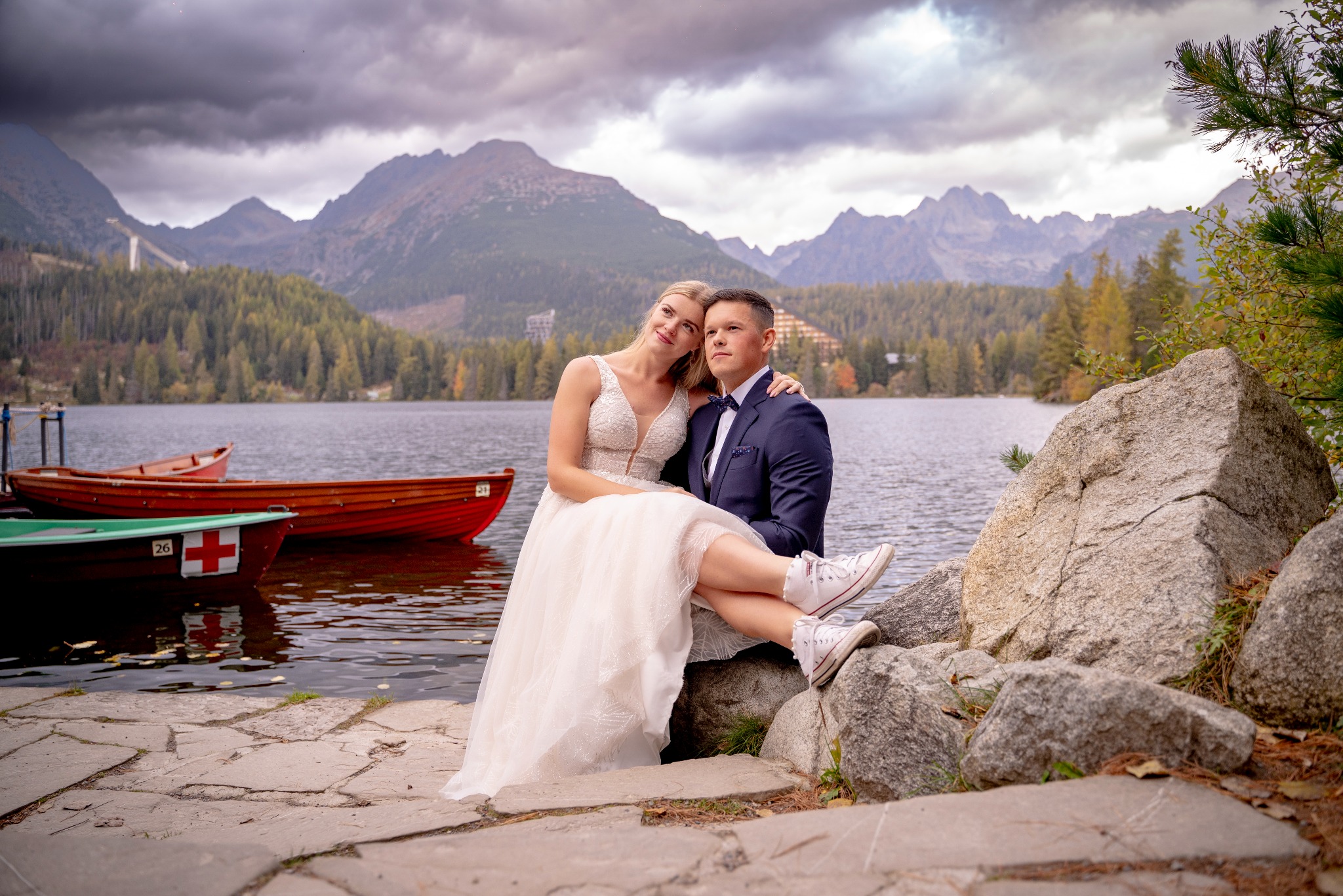 Para młoda w strojach ślubnych siedzi na kamieniach nad jeziorem z łódkami, góry w tle, panna młoda ma na nogach białe trampki.