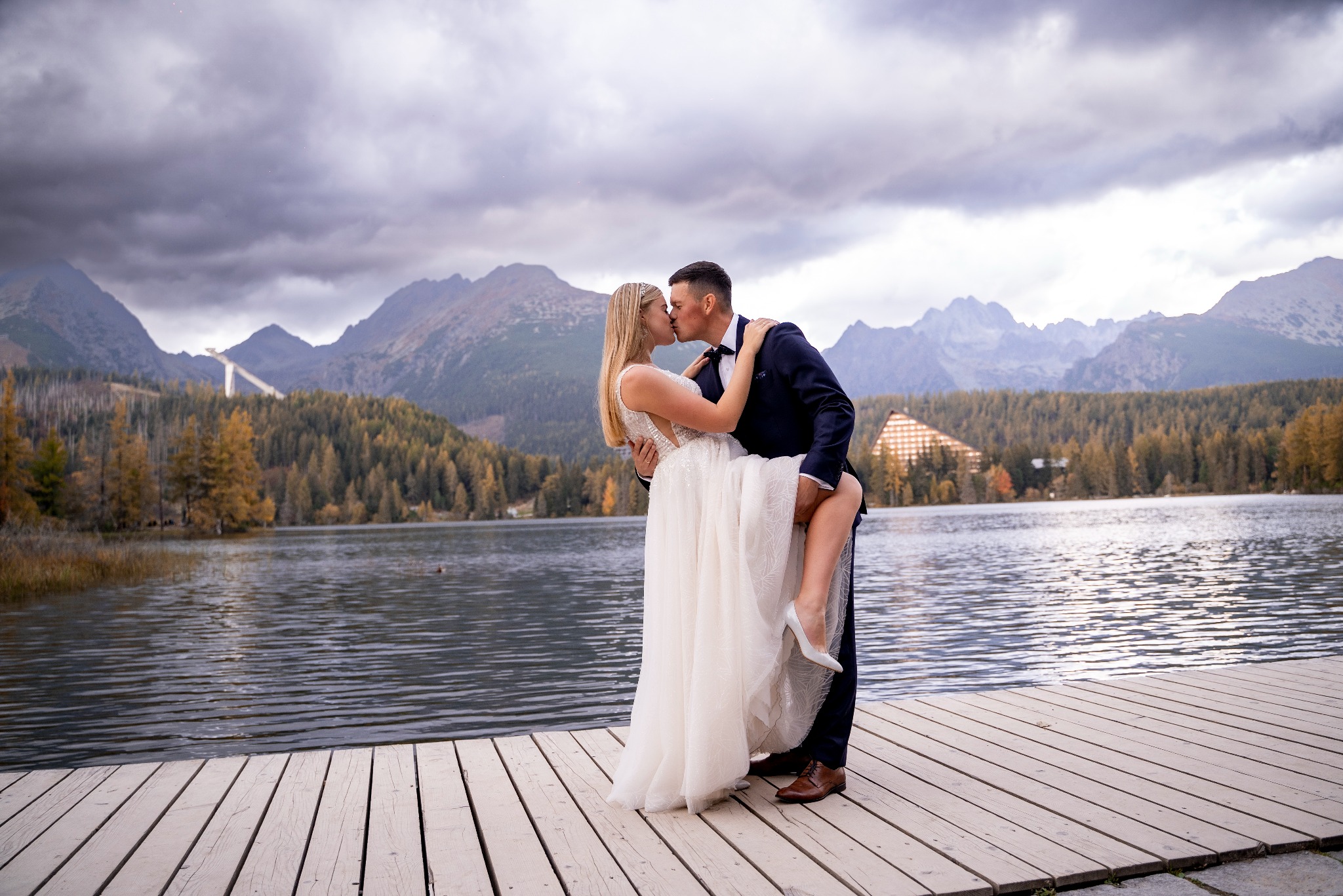 Panna młoda w białej sukni i pan młody w granatowym garniturze całują się na drewnianym pomoście nad jeziorem, z górami w tle i pochmurnym niebem.