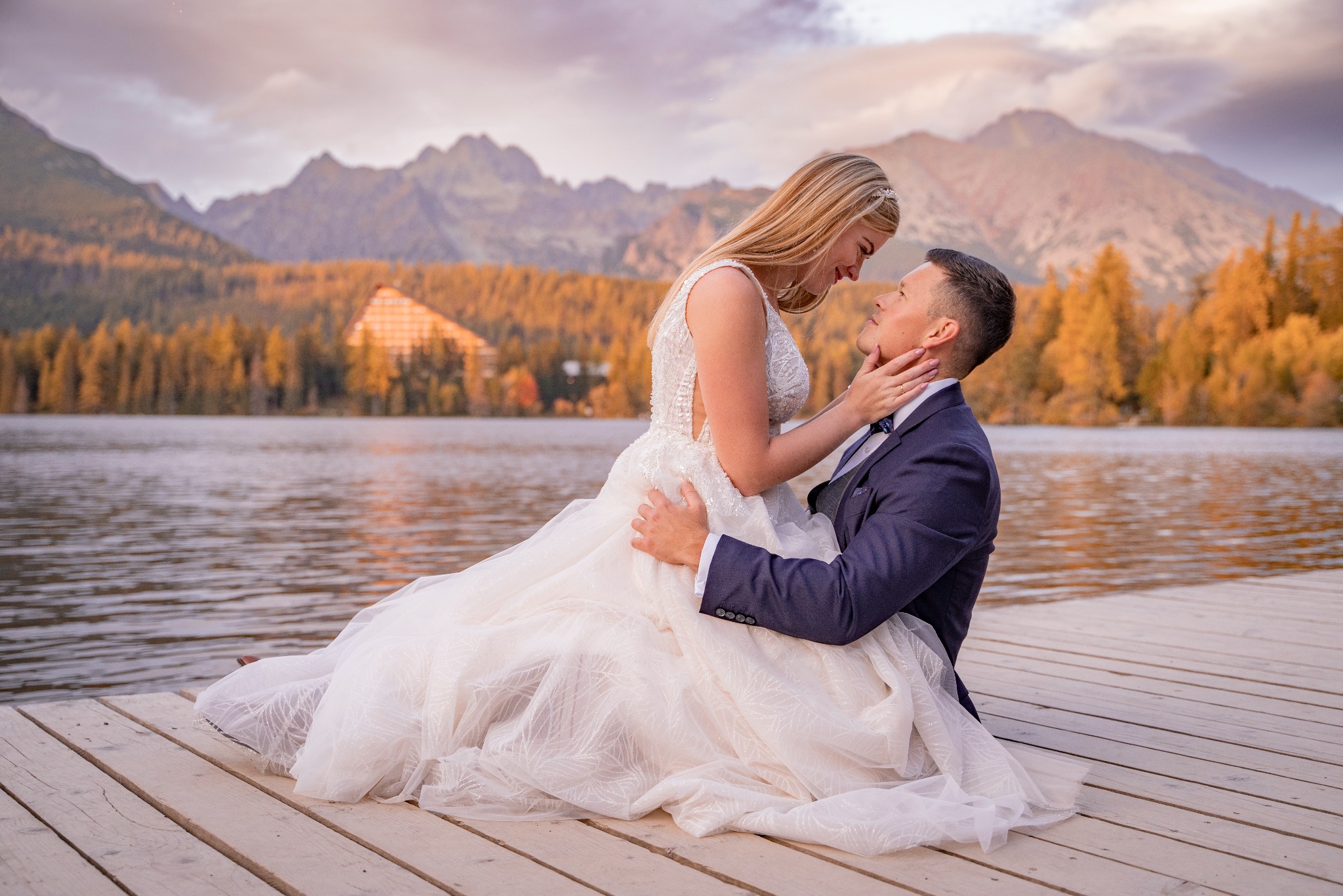 Para młoda siedzi na drewnianym pomoście nad jeziorem, w tle góry i jesienny las. Panna młoda w białej sukni z koronkową górą, pan młody w granatowym garniturze z muchą. Delikatne oświetlenie...