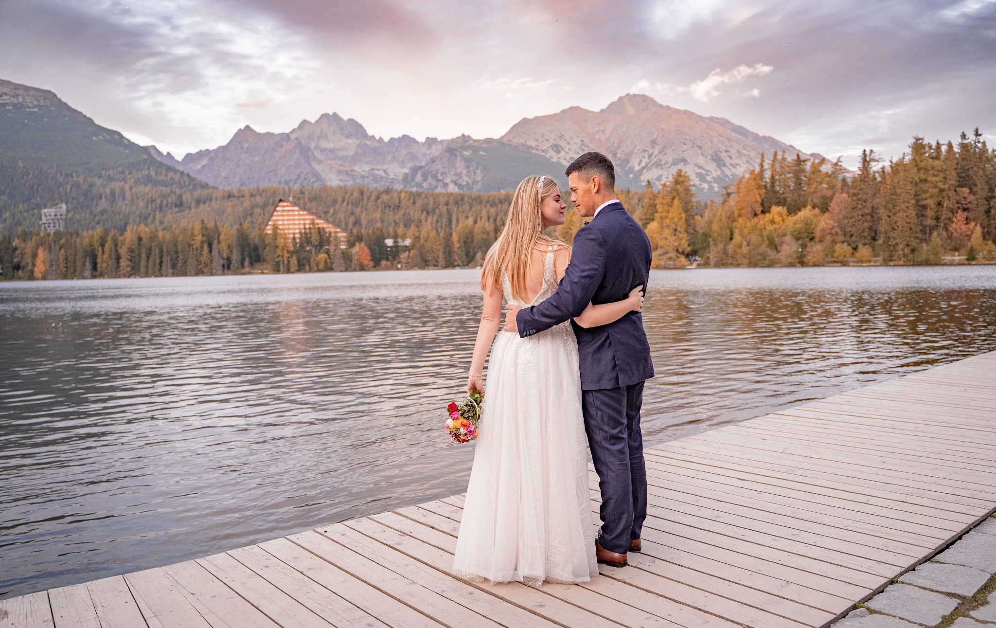 Para młoda obejmująca się na drewnianym pomoście nad jeziorem z widokiem na góry w tle. Panna młoda w białej sukni z odkrytymi plecami trzyma bukiet kwiatów.