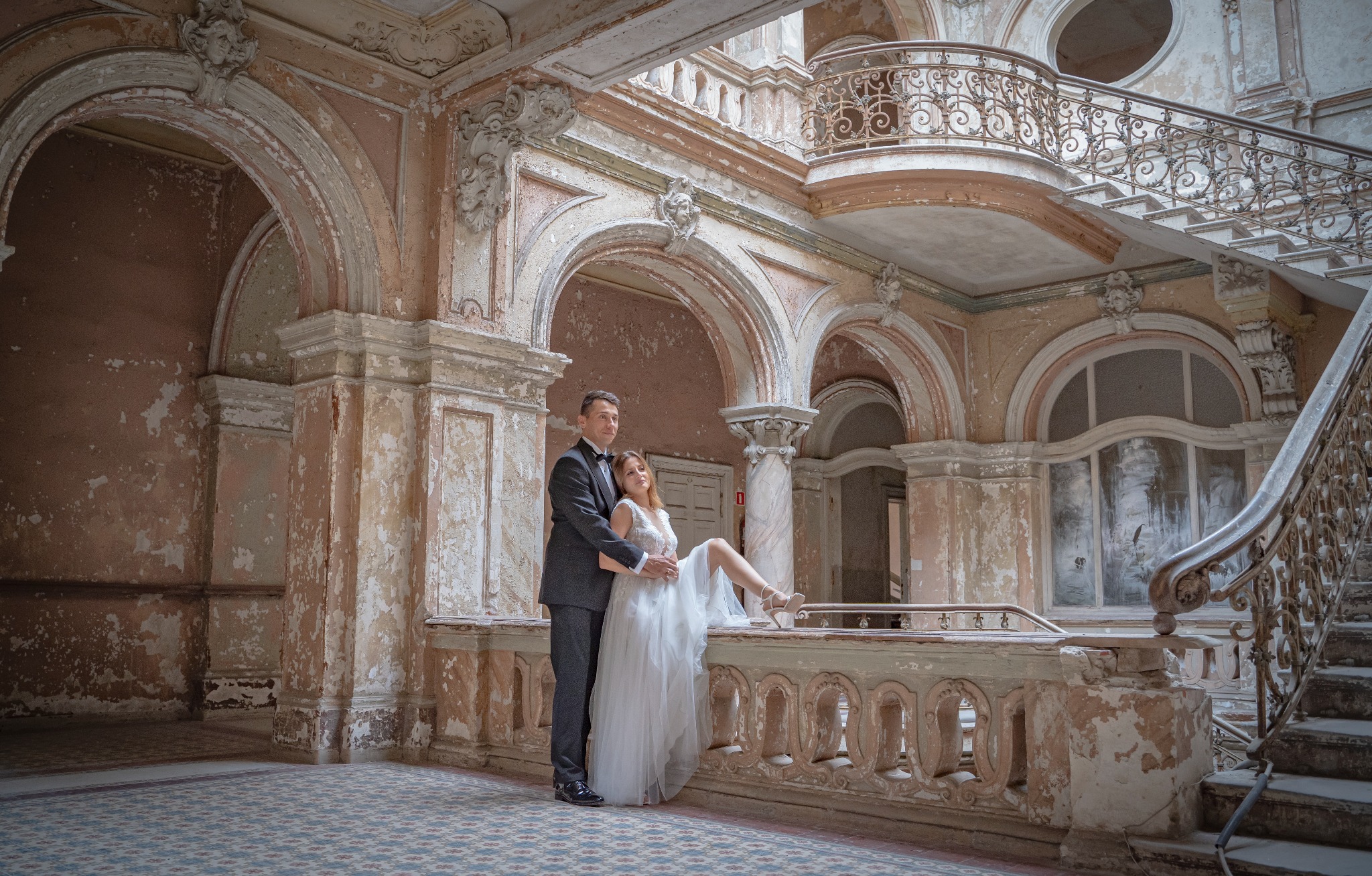 Para młoda: mężczyzna w ciemnym garniturze i kobieta w białej sukni ślubnej pozują w opuszczonym pałacu z dekoracyjnymi łukami, schodami i balkonem z kutą balustradą.