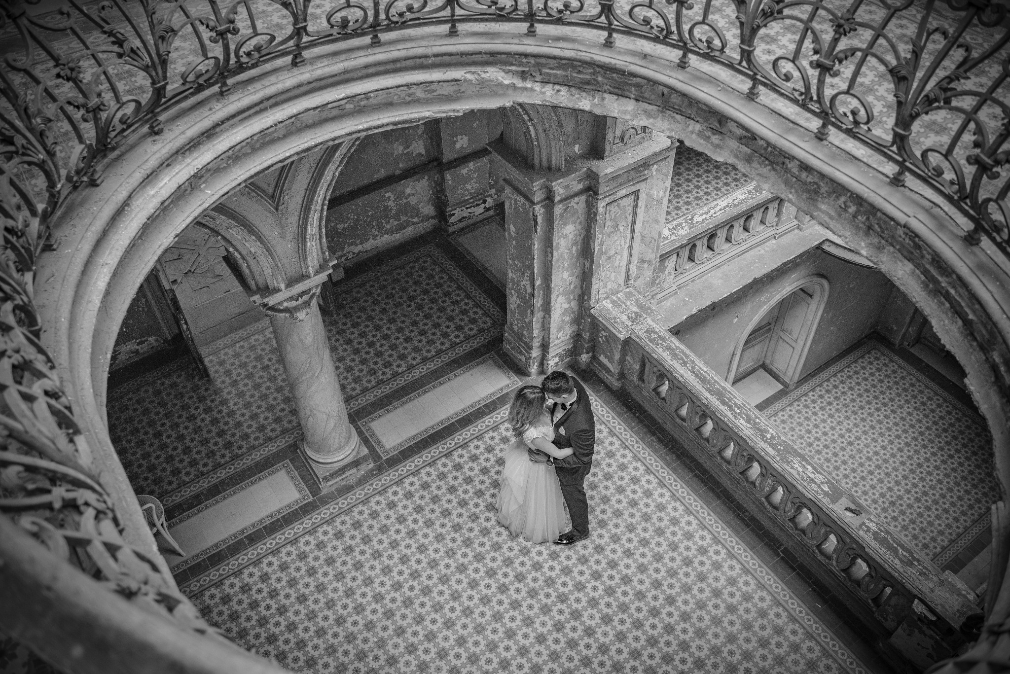 Czarno-białe ujęcie pary młodej obejmującej się na wzorzystej posadzce w zabytkowym wnętrzu, widok z góry zza ozdobnej balustrady.