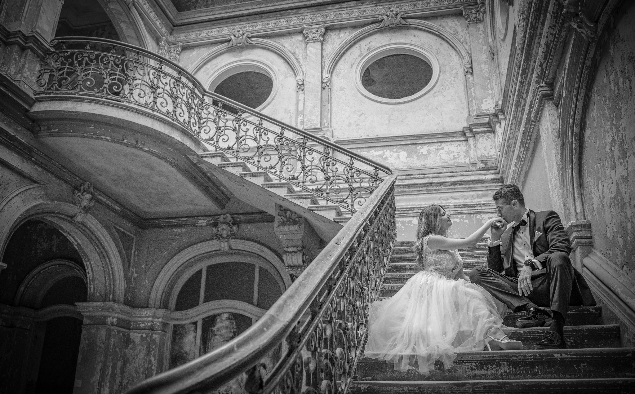 Czarno-białe zdjęcie pary: kobieta w sukni ślubnej całuje dłoń mężczyzny w smokingu, siedzą na schodach w opuszczonym budynku z dekoracyjnymi balustradami i okrągłymi oknami.