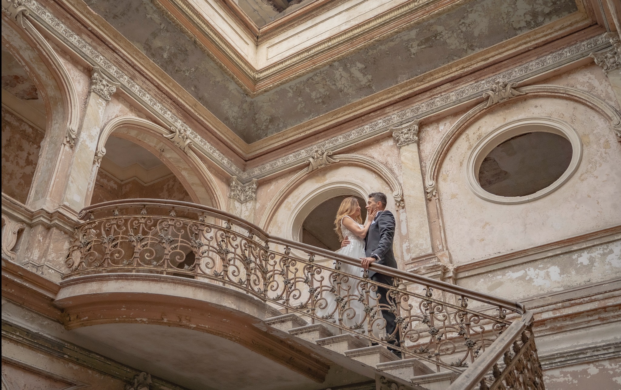 Para młoda na schodach z kutą balustradą w opuszczonym pałacu z łuszczącą się farbą i dekoracyjnymi łukami.