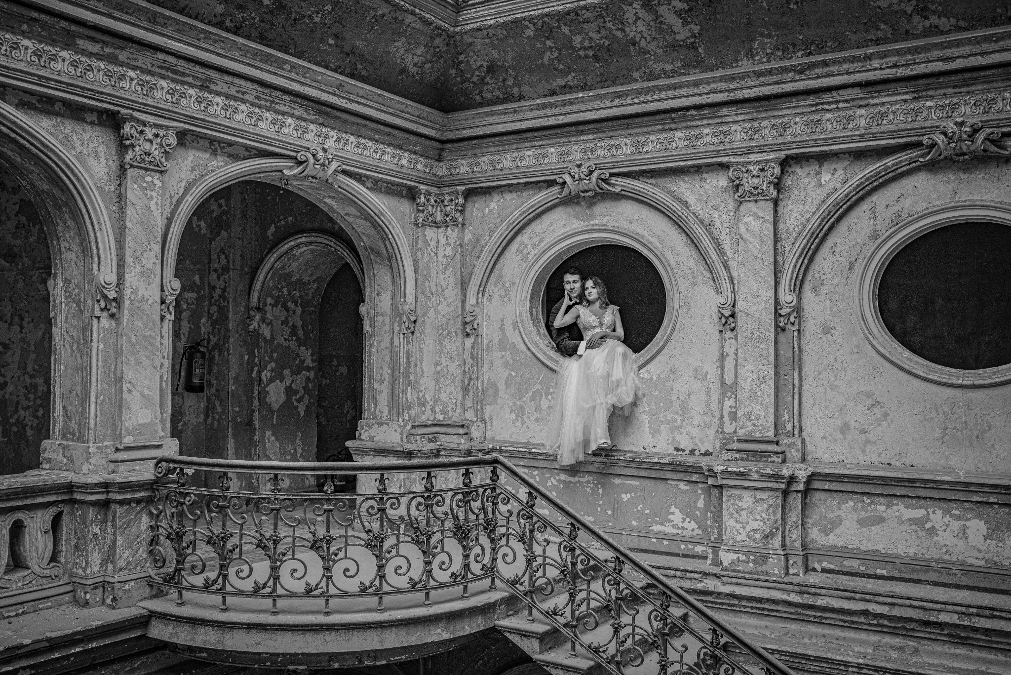 Para młoda w czerni i bieli pozuje w okrągłym oknie pałacu z dekoracyjnymi elementami architektonicznymi i zniszczonymi ścianami.