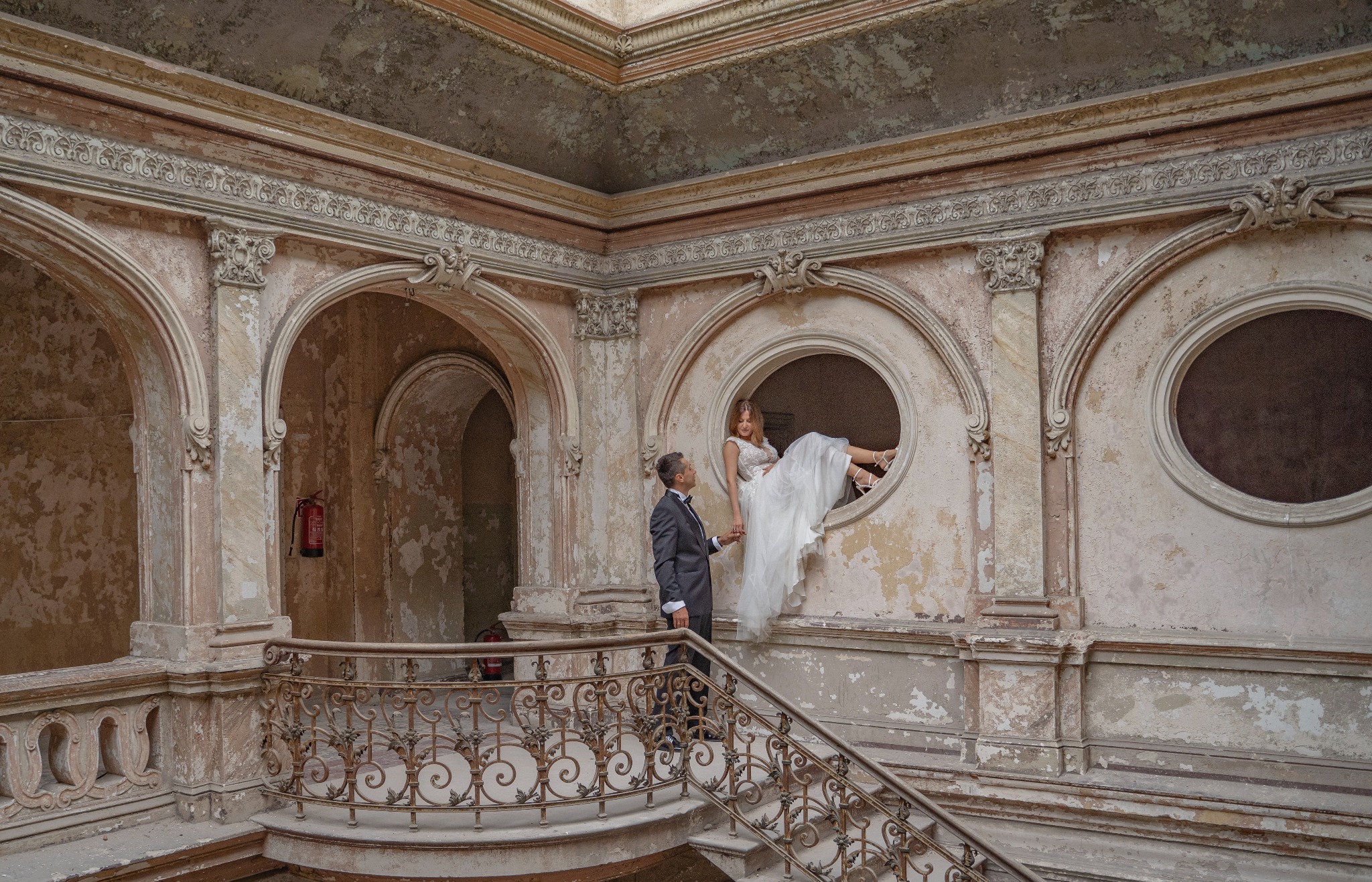 Para młoda w Bytomiu pozuje w opuszczonym pałacu, panna młoda siedzi w okrągłym oknie, pan młody stoi na schodach i trzyma ją za rękę.