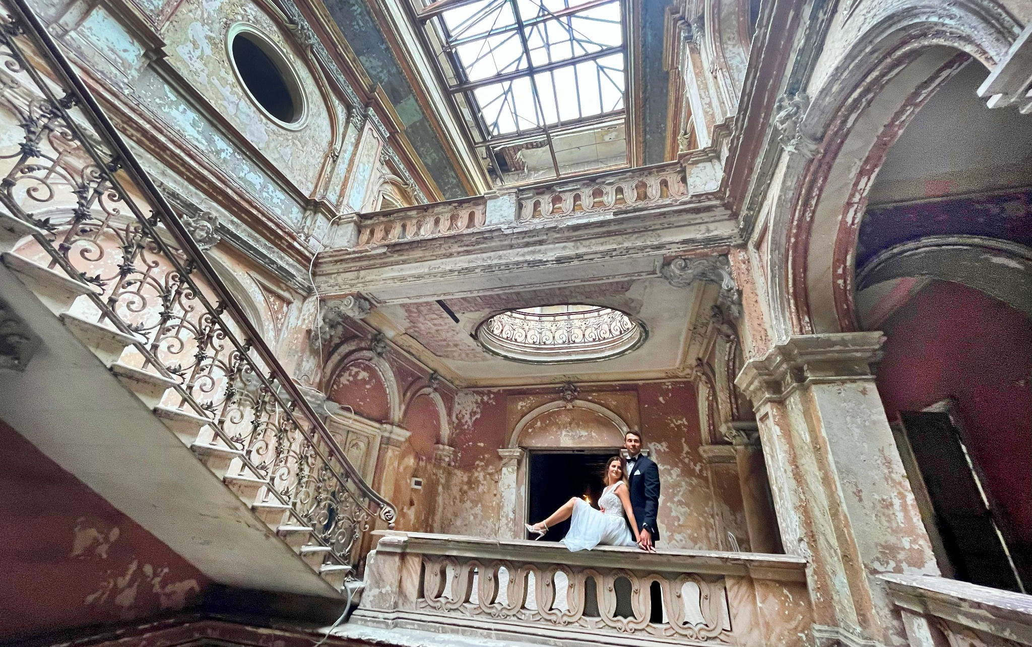 Para młoda siedząca na balustradzie w opuszczonym pałacu z widocznymi schodami, balkonem i świetlikiem w dachu.