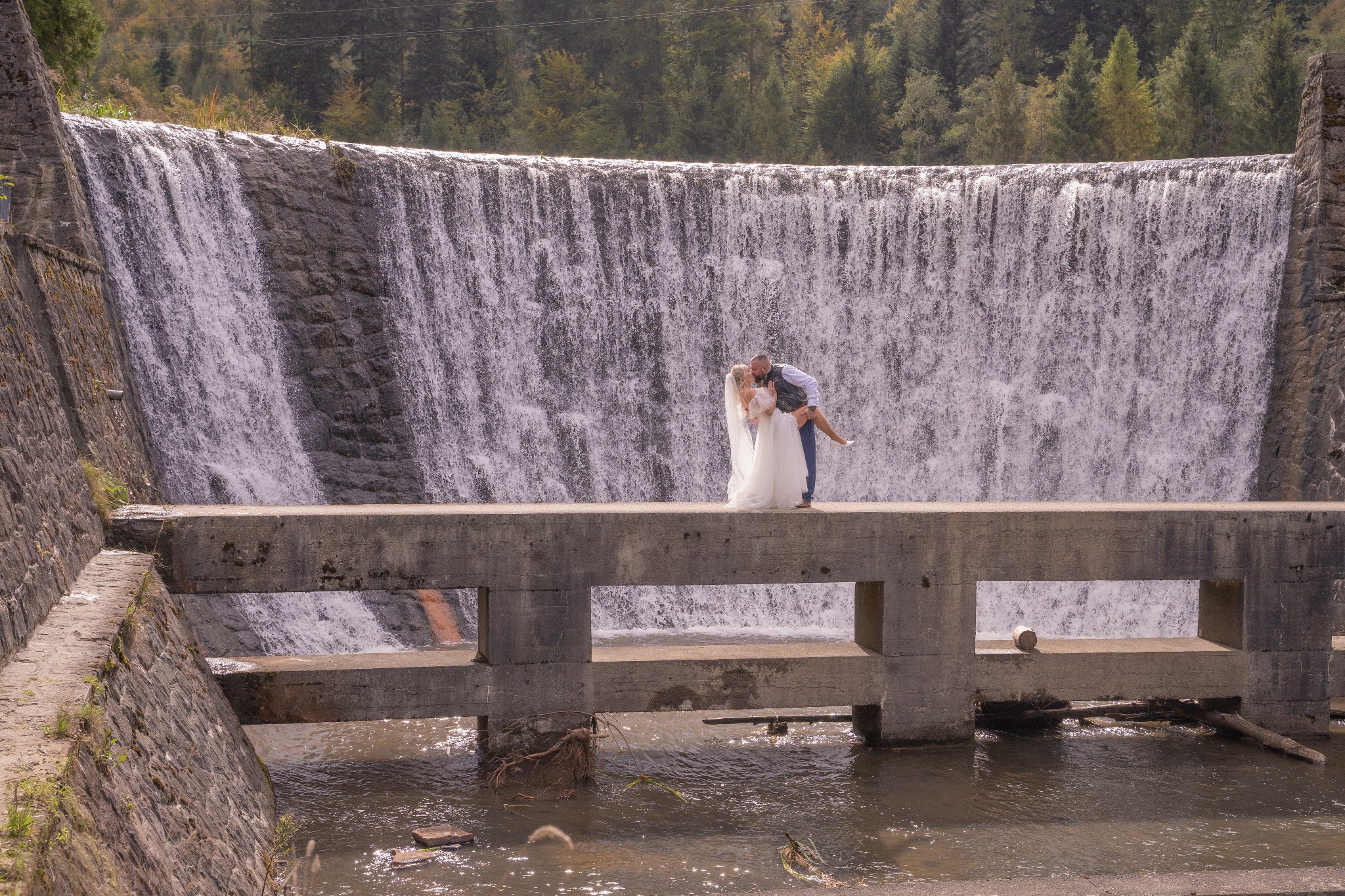 Para młoda na moście przed wodospadem: Panna Młoda w białej sukni ślubnej z welonem, Pan Młody w kamizelce i jeansach, podnosi ją na rękach. W tle kamienny wodospad i drzewa.