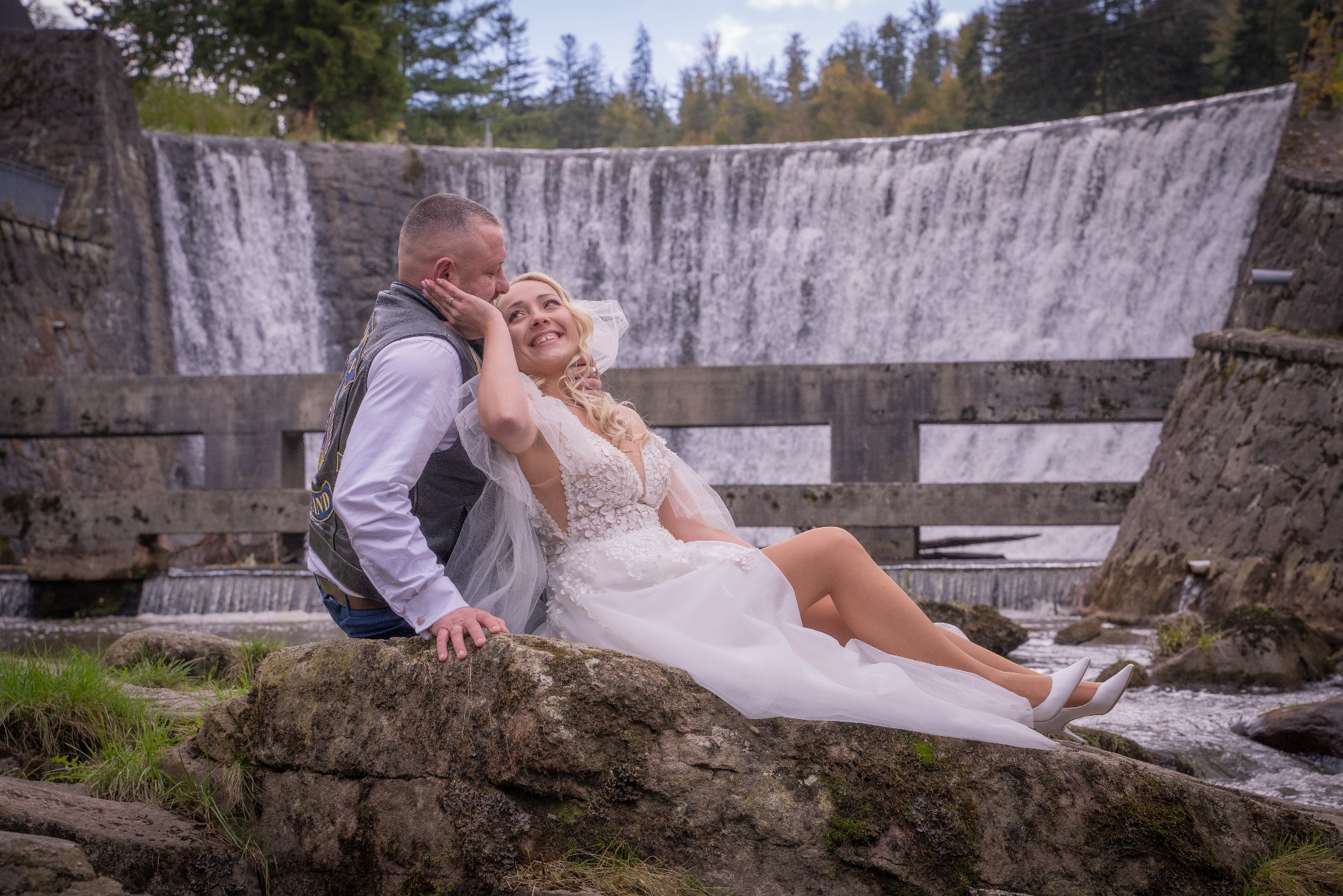 Para młoda w sesji ślubnej siedzi na kamieniu przed wodospadem, panna młoda w sukni z koronkowymi aplikacjami i welonem, pan młody w kamizelce motocyklowej.