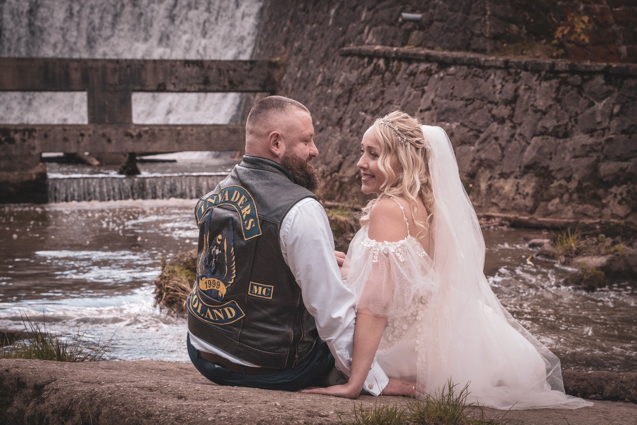 Para młoda siedzi na kamieniu przed wodospadem; panna młoda w zwiewnej sukni i welonie, pan młody w skórzanej kamizelce motocyklowej z naszywkami, oboje uśmiechnięci.