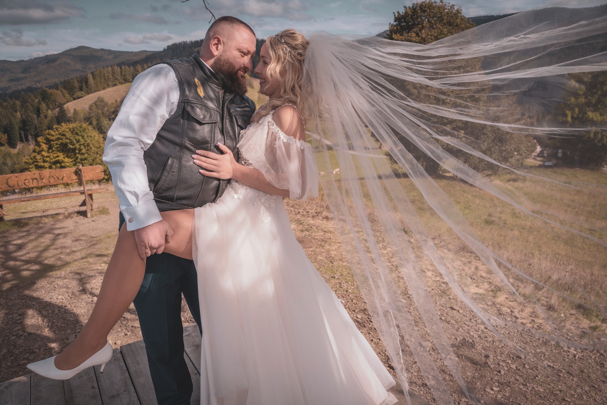 Para młoda w malowniczej scenerii górskiej: mężczyzna w skórzanej kamizelce trzyma pannę młodą w białej sukni i welonie, który powiewa na wietrze.