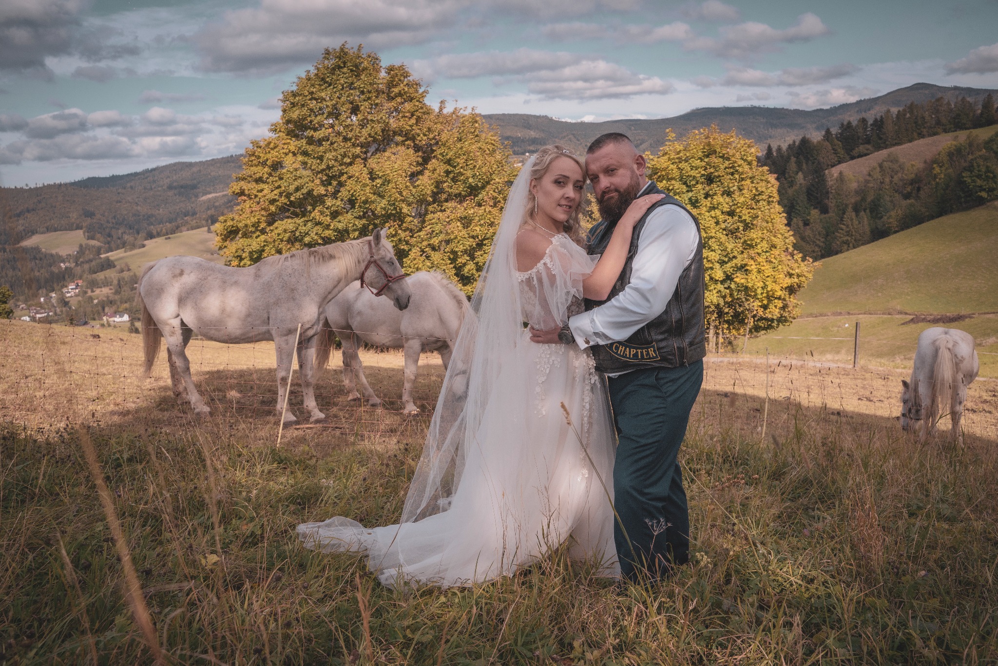 Para młoda pozuje w plenerze z białymi końmi na tle gór, panna młoda w białej sukni z welonem, pan młody w skórzanej kamizelce.