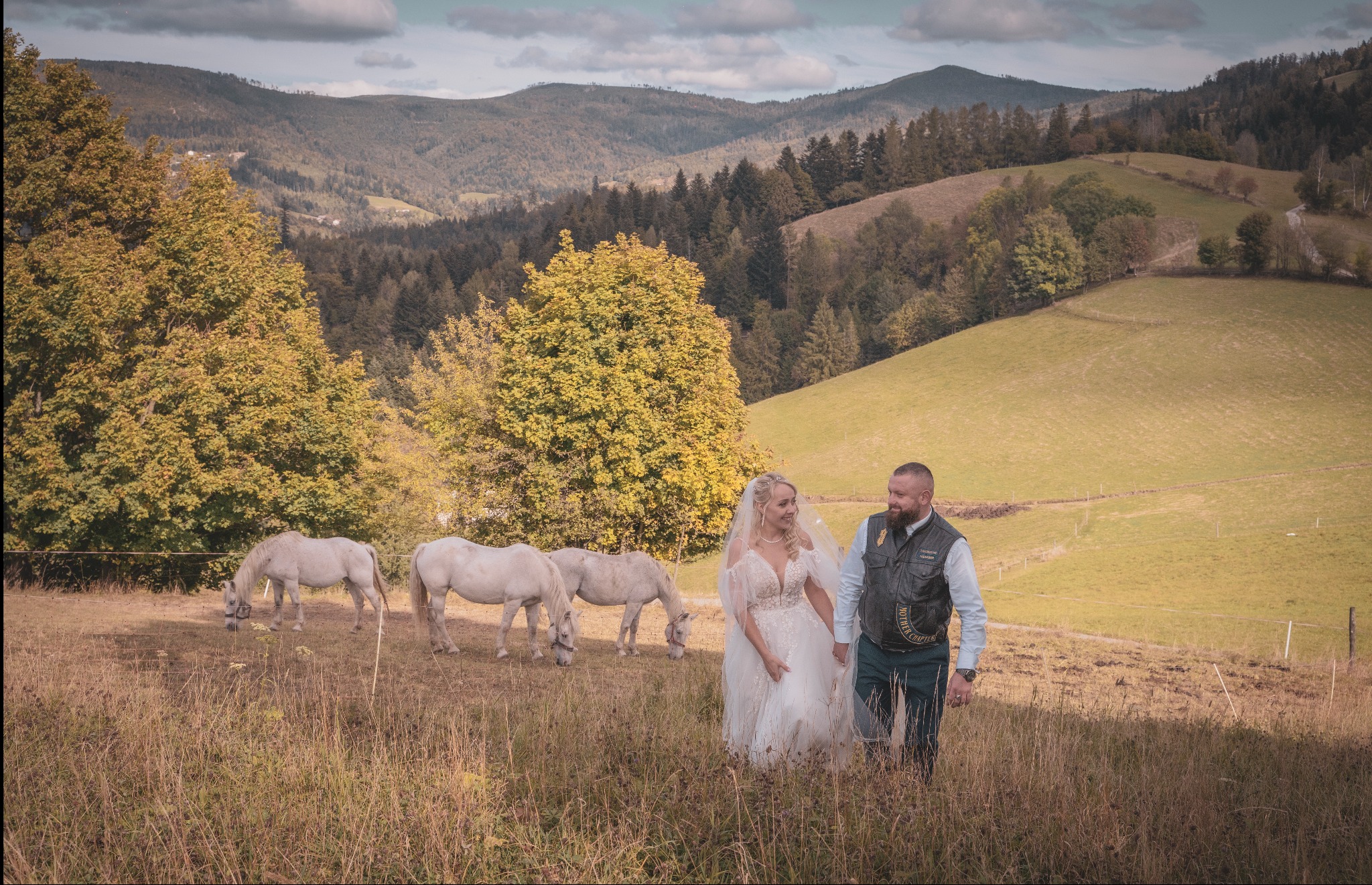Para młoda trzymająca się za ręce spaceruje po łące z białymi końmi na tle malowniczego krajobrazu górskiego.